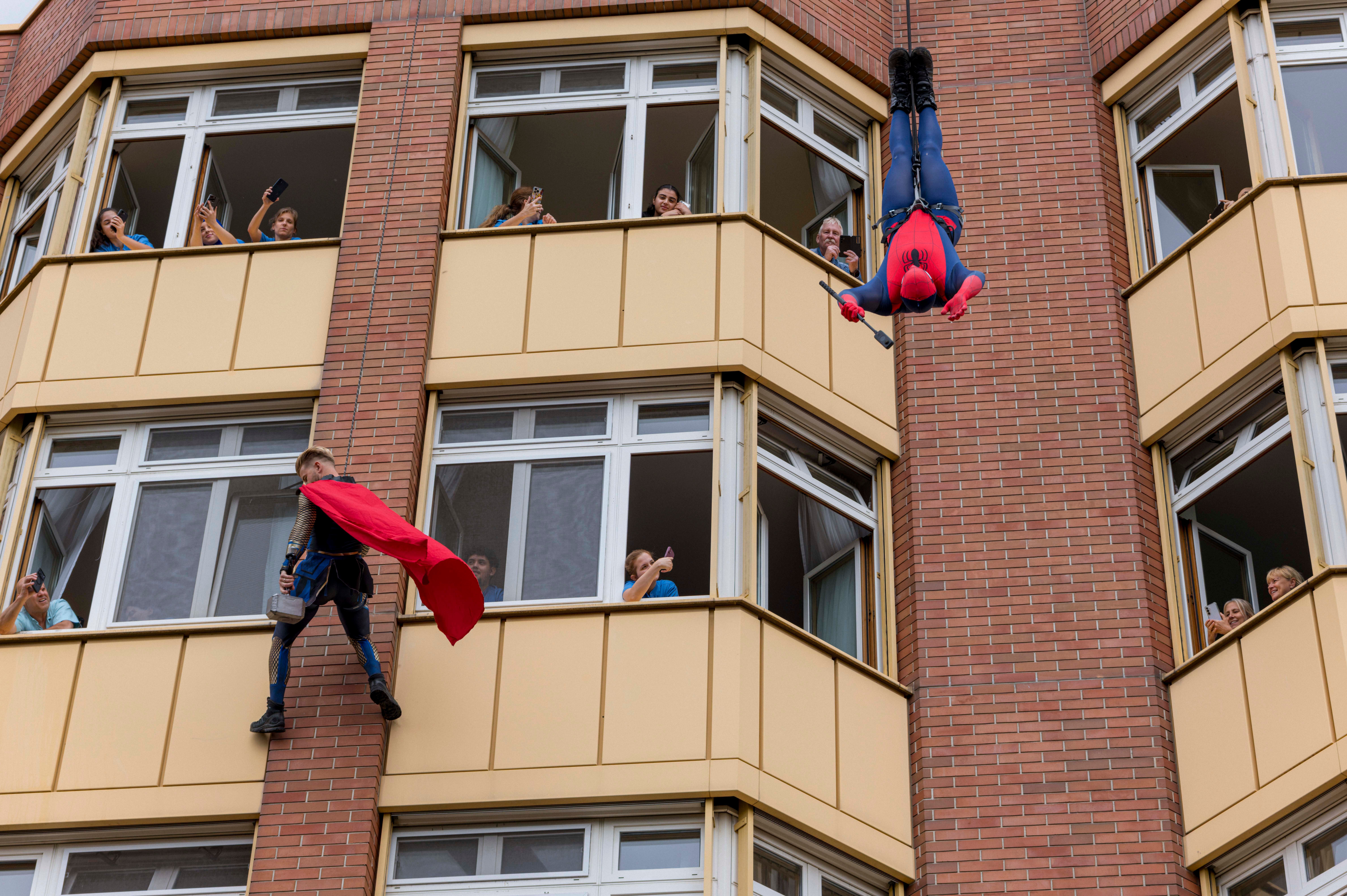 Zwei als Superhelden verkleidete Personen klettern an einem Haus, Kinder sehen begeistert aus den Fenstern.