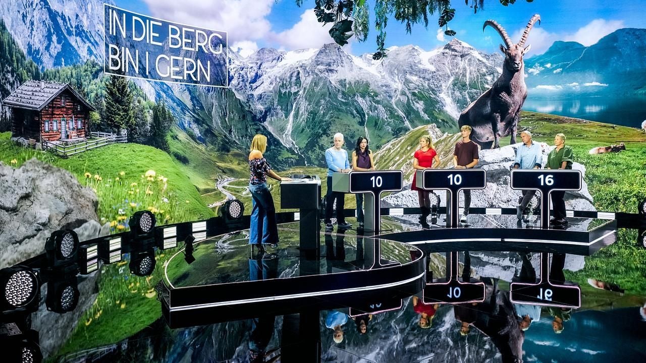 Quizshow mit fünf Kandidaten vor Alpenlandschaft und der Aufschrift "IN DIE BERG BIN I GERN".
