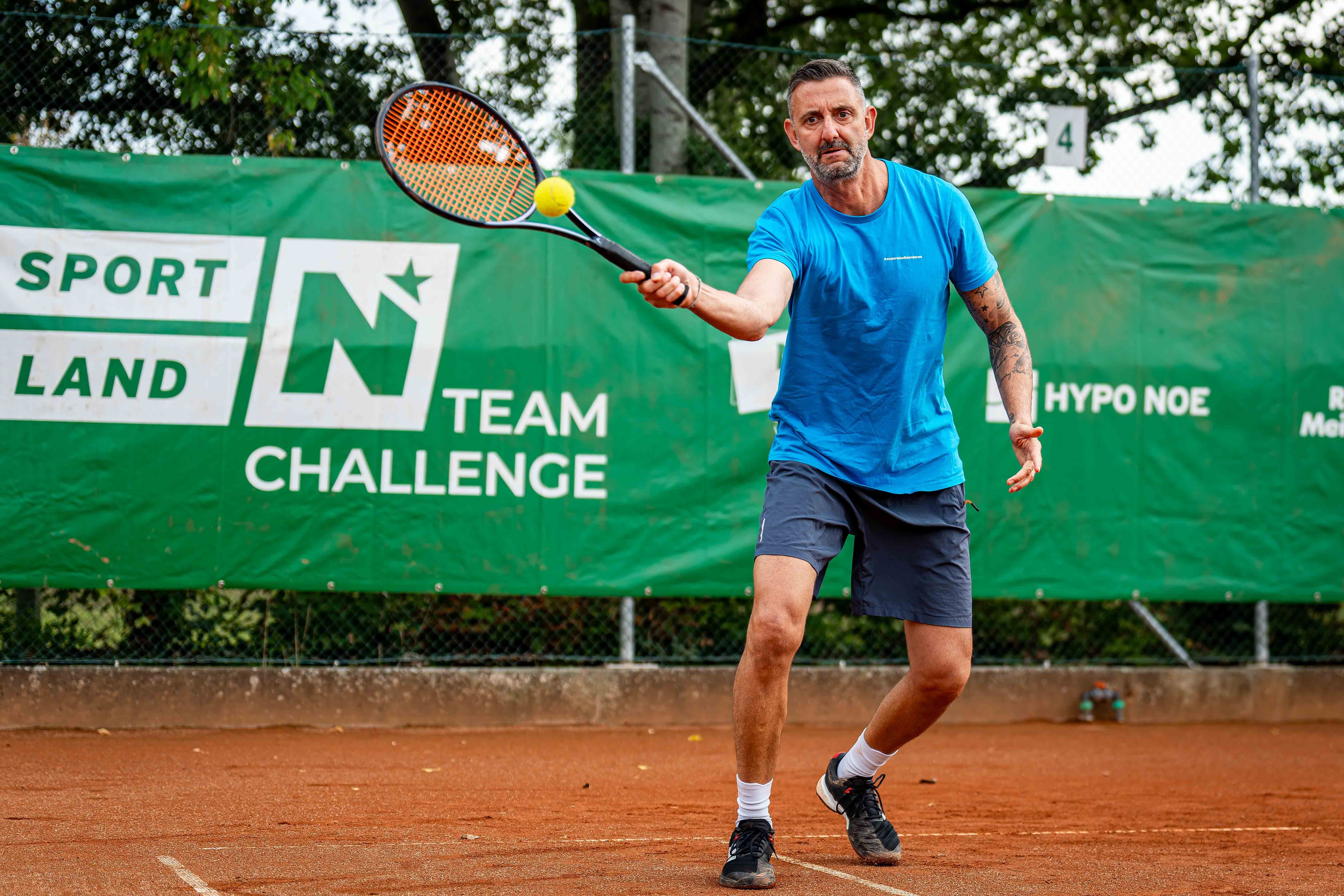 Mann in blauer Sportkleidung spielt Tennis auf einem Sandplatz vor grünem Turnierbanner.
