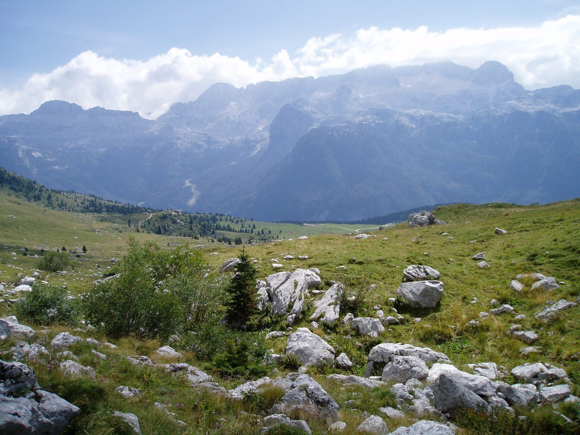 Grüne Bergwiese mit Felsen vor bewölkten, hohen Kaninbergen im Hintergrund.
