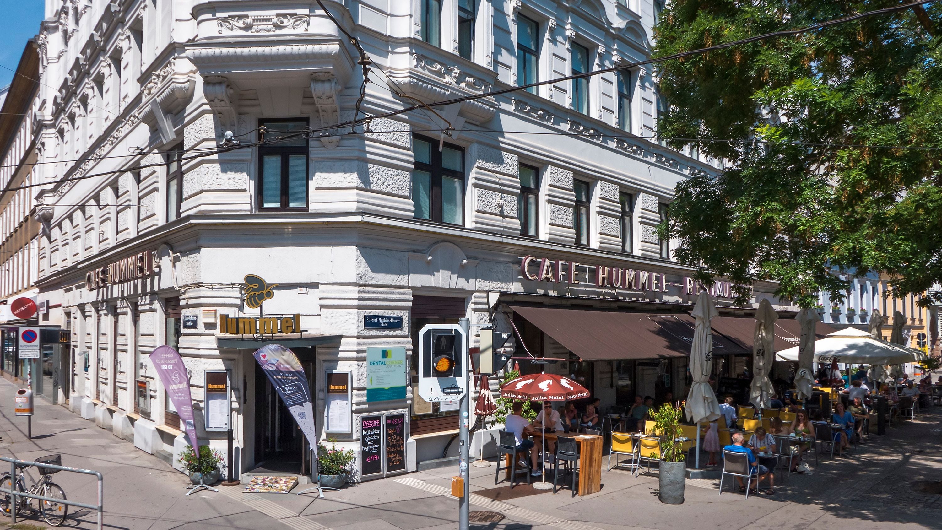 Straßencafé mit Gästen unter Sonnenschirmen vor dem historischen Gebäude des Café Hummel.