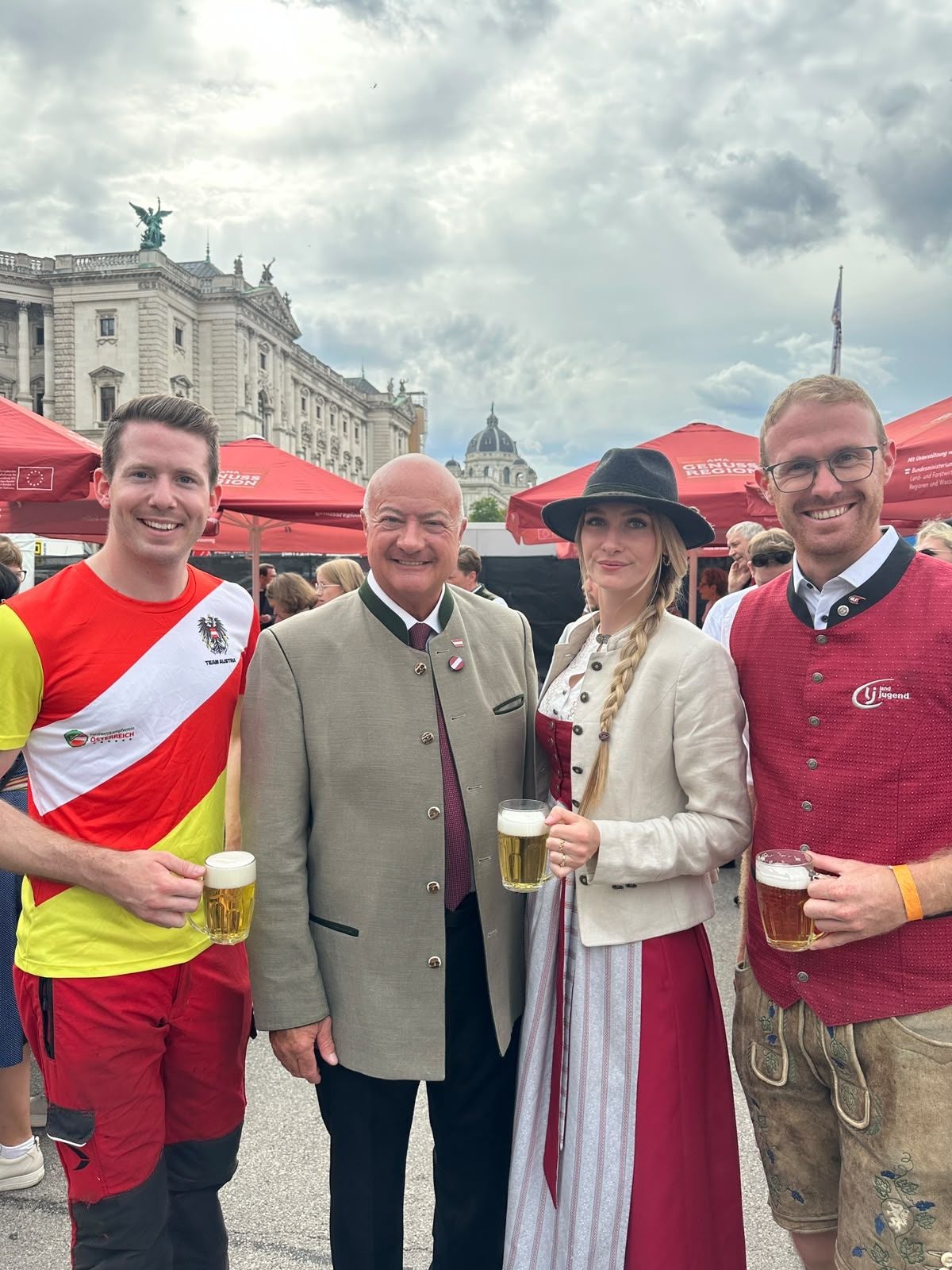 Vier Personen in Tracht und Sportkleidung mit Bierkrügen auf einem Fest vor historischen Gebäuden.