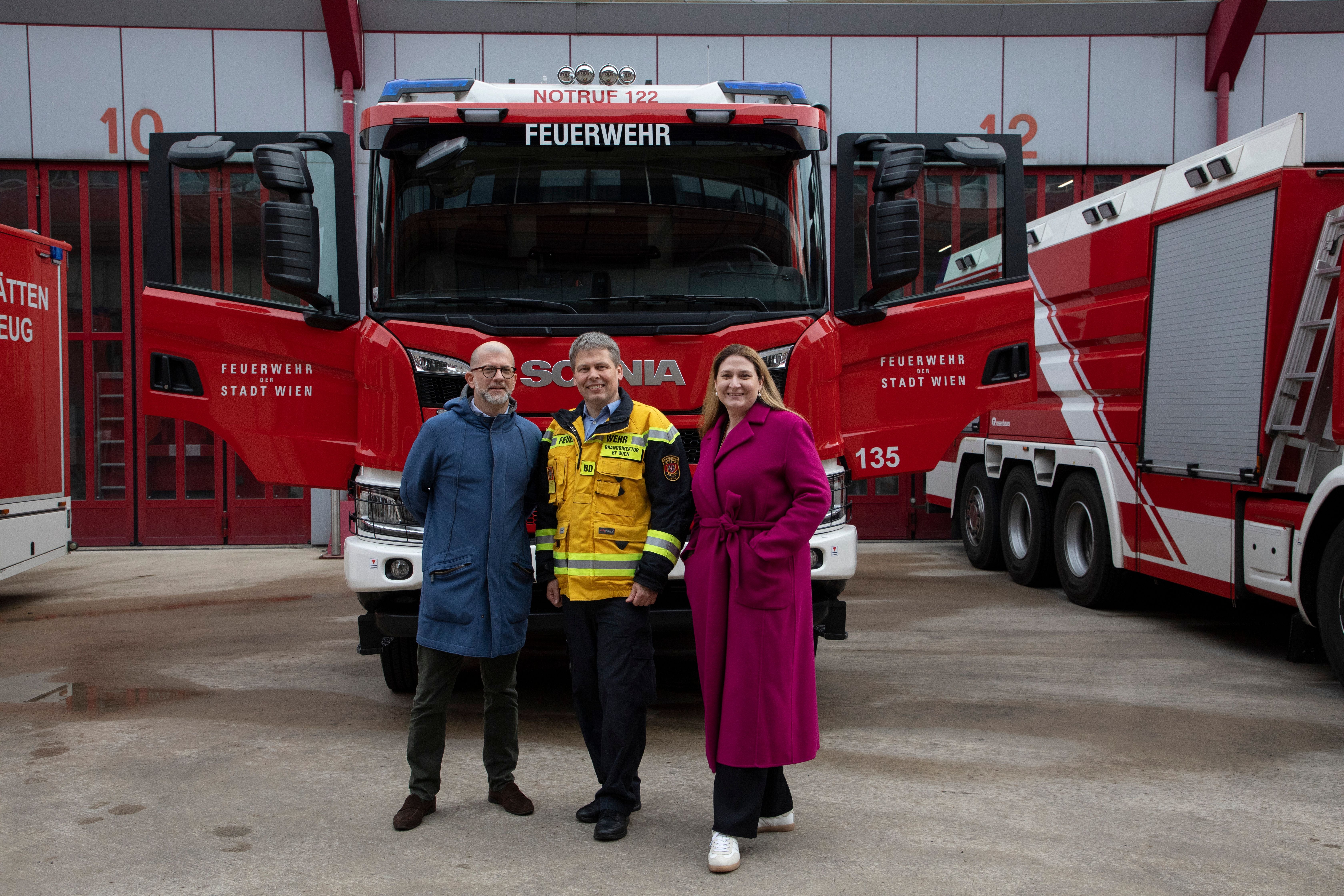 Drei Personen stehen vor einem Feuerwehrfahrzeug der Stadt Wien mit geöffneten Türen.