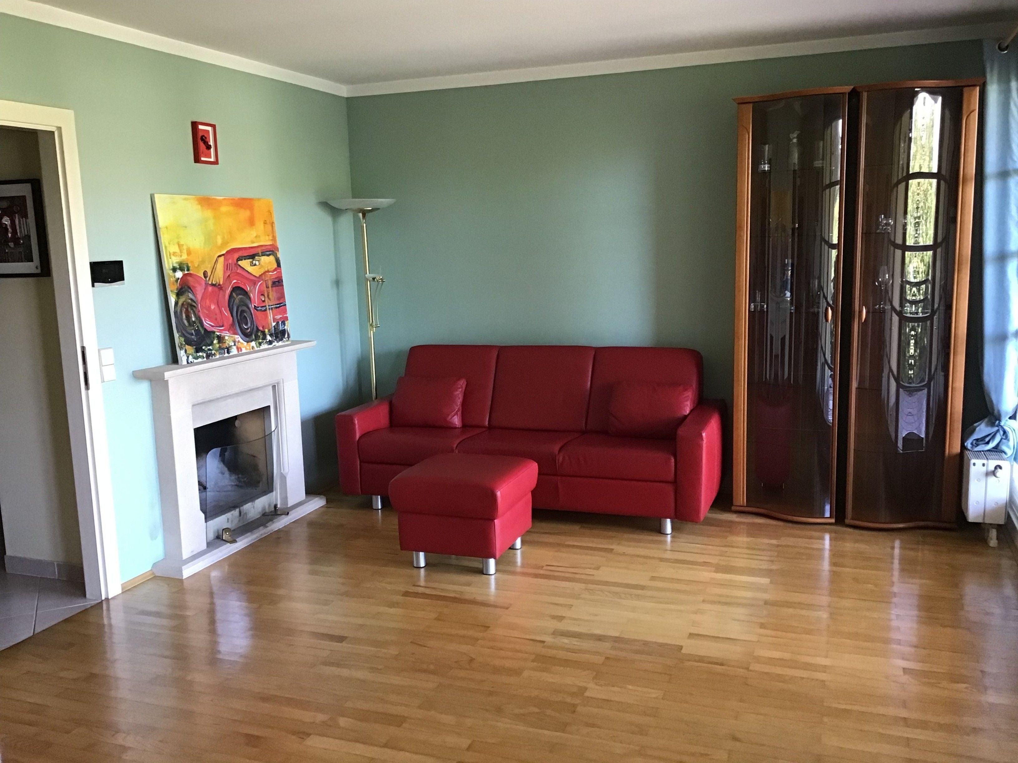 Wohnzimmer mit rotem Sofa, Kamin, Gemälde, Vitrine und Parkettboden.