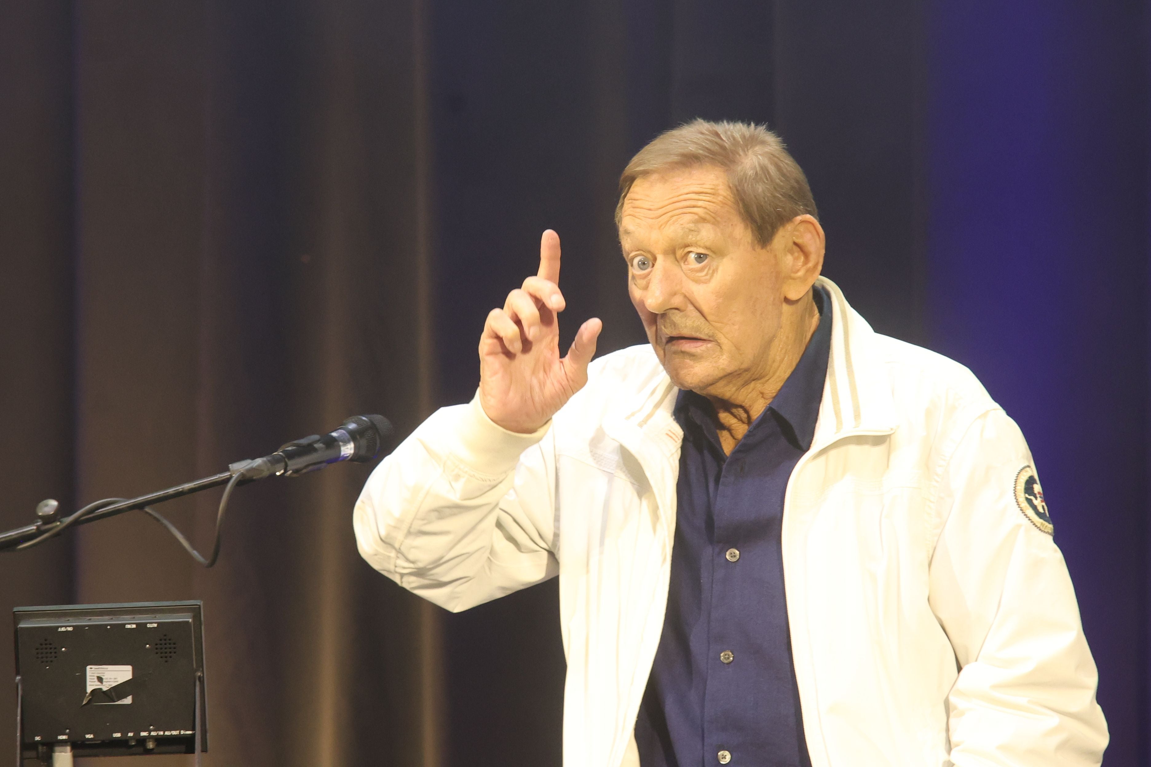 Ein älterer Mann spricht auf einer Bühne und hebt nachdenklich den Zeigefinger.