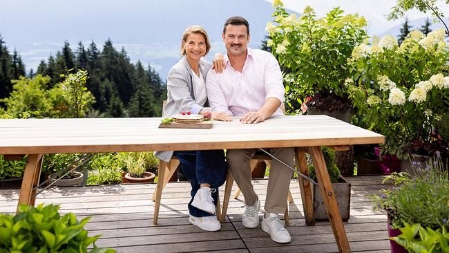 Ein Paar sitzt lächelnd an einem Holztisch auf einer Terrasse mit Blumen und Blick auf Berge.