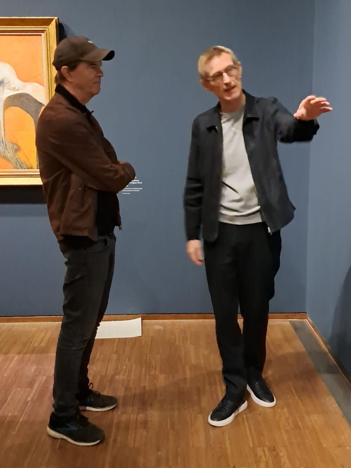 Zwei Männer unterhalten sich in einem Museum vor einem blauen Hintergrund und einem Gemälde.