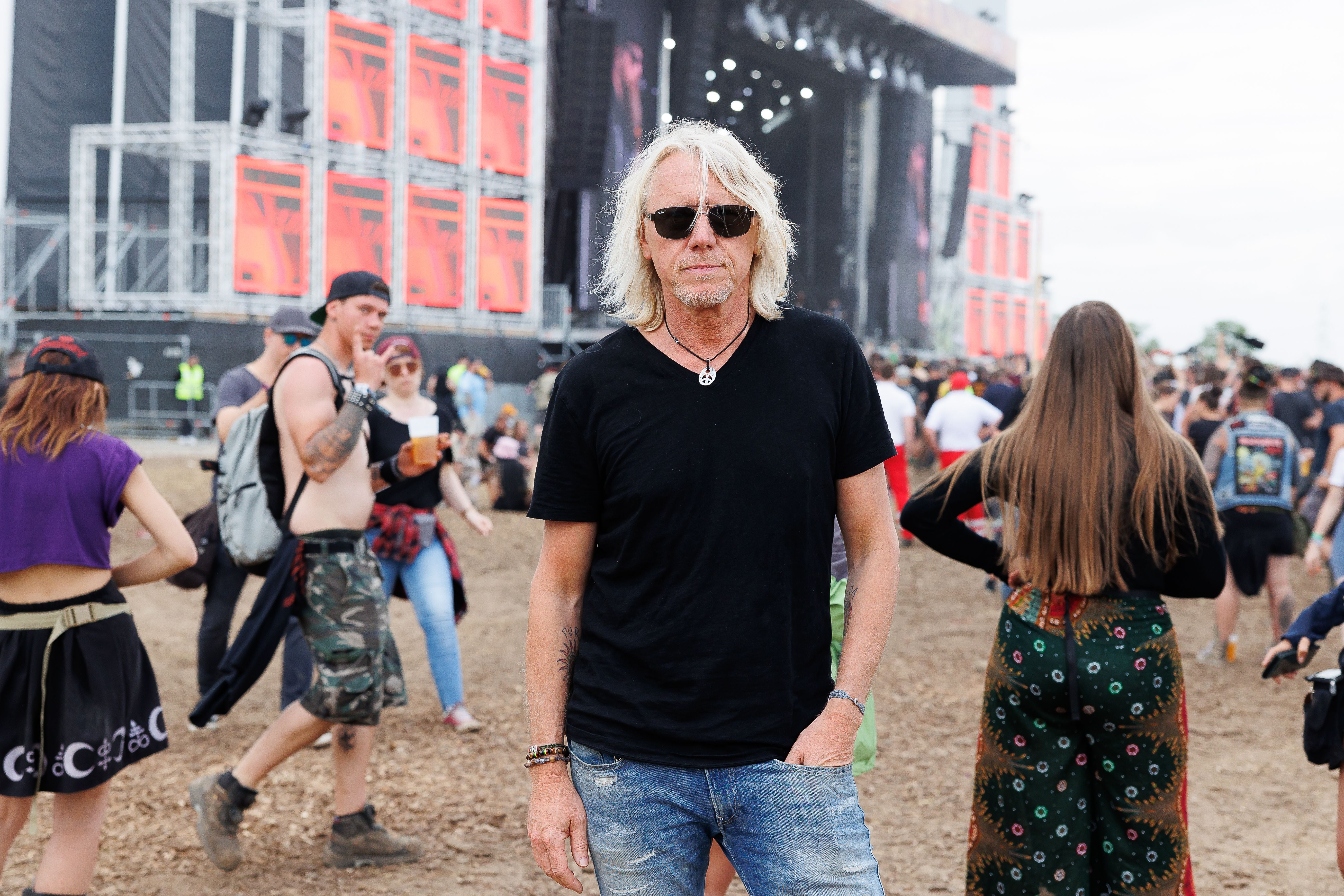Mann mit Sonnenbrille steht auf einem Musikfestivalgelände, Menschen und Bühne im Hintergrund.