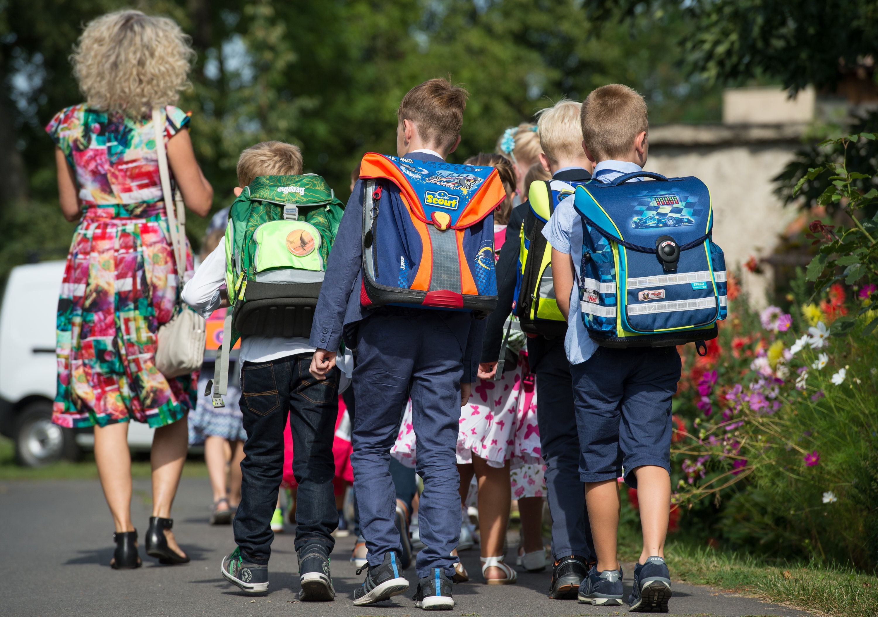 Mehrere Grundschulkinder mit bunten Schulranzen gehen gemeinsam mit einer Frau zur Schule.