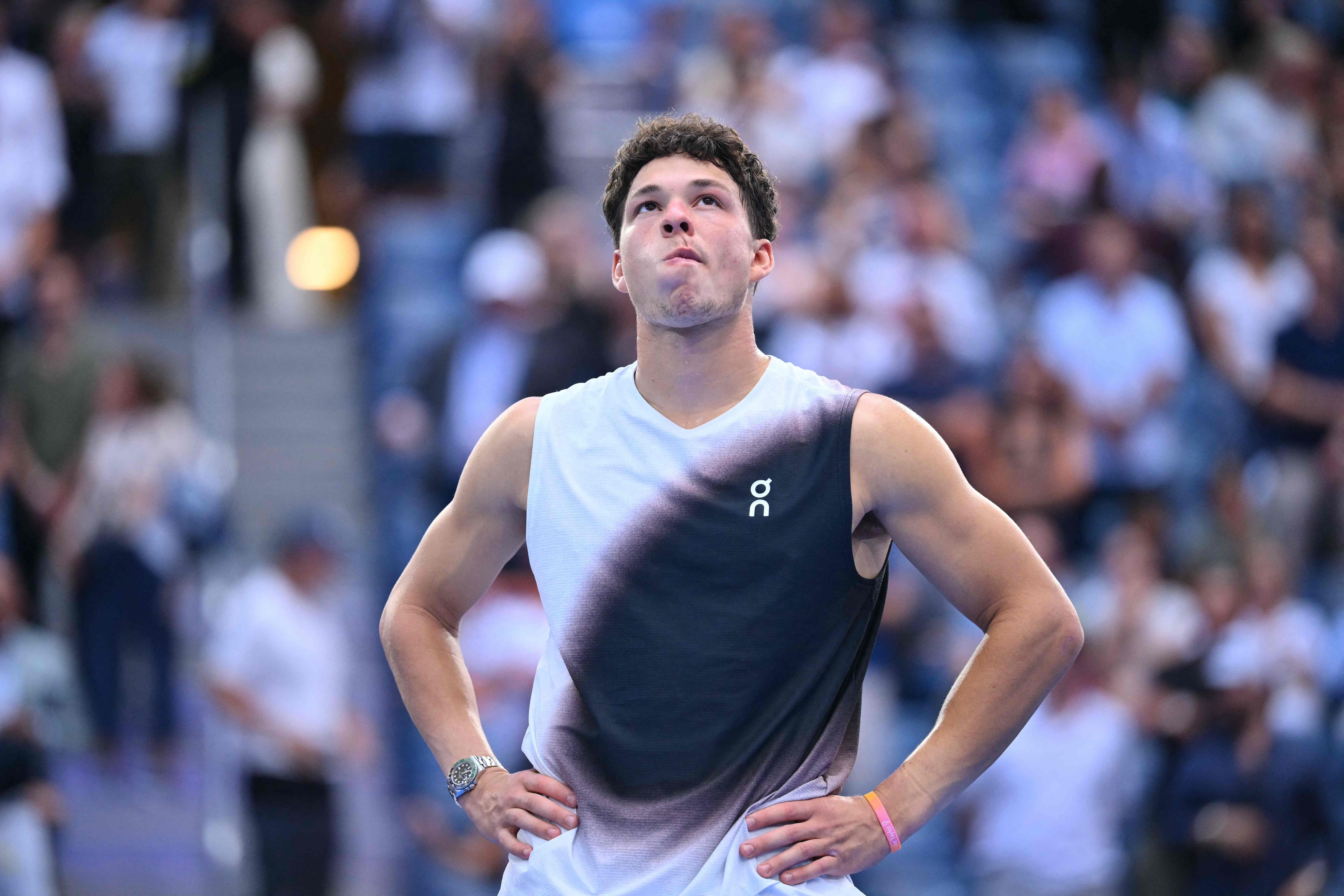 Ein Tennisspieler steht nachdenklich mit Händen in der Hüfte auf dem Platz, Zuschauer im Hintergrund.
