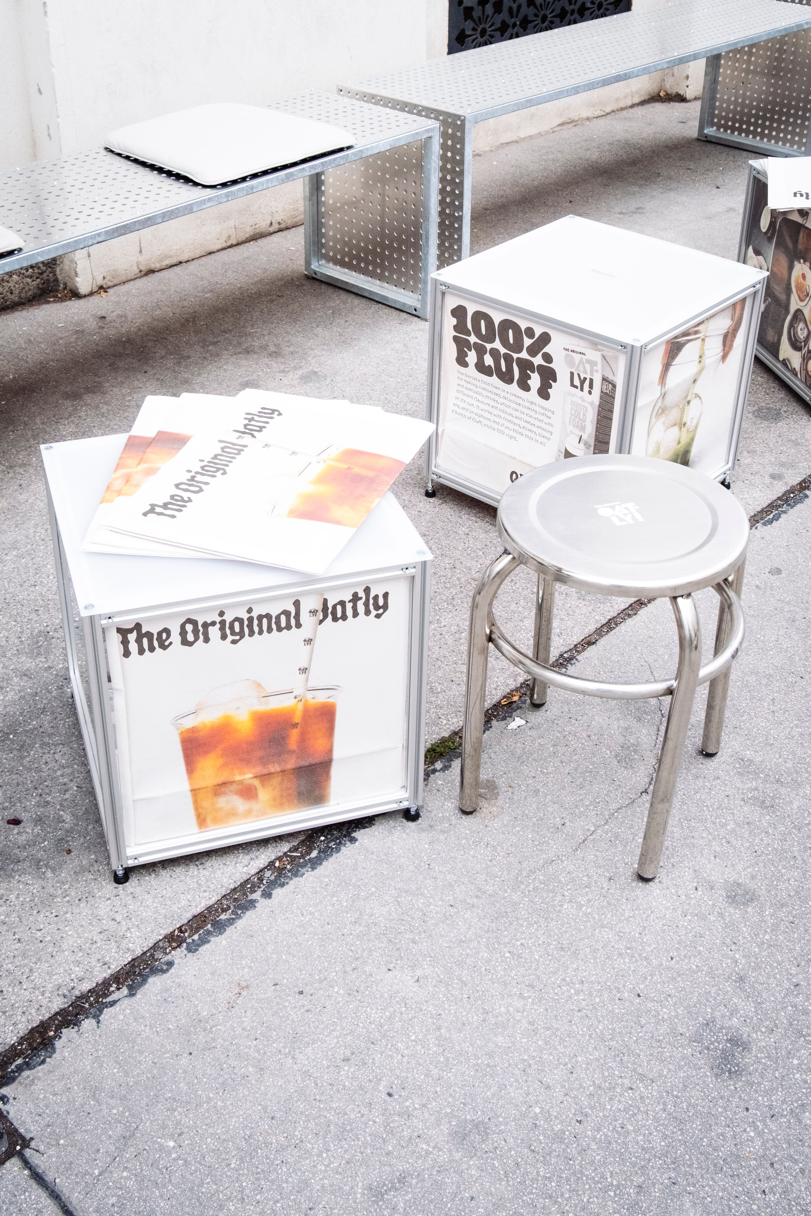 Metallhocker, Sitzwürfel mit Oatly-Werbung und Zeitungen stehen auf dem Gehweg.