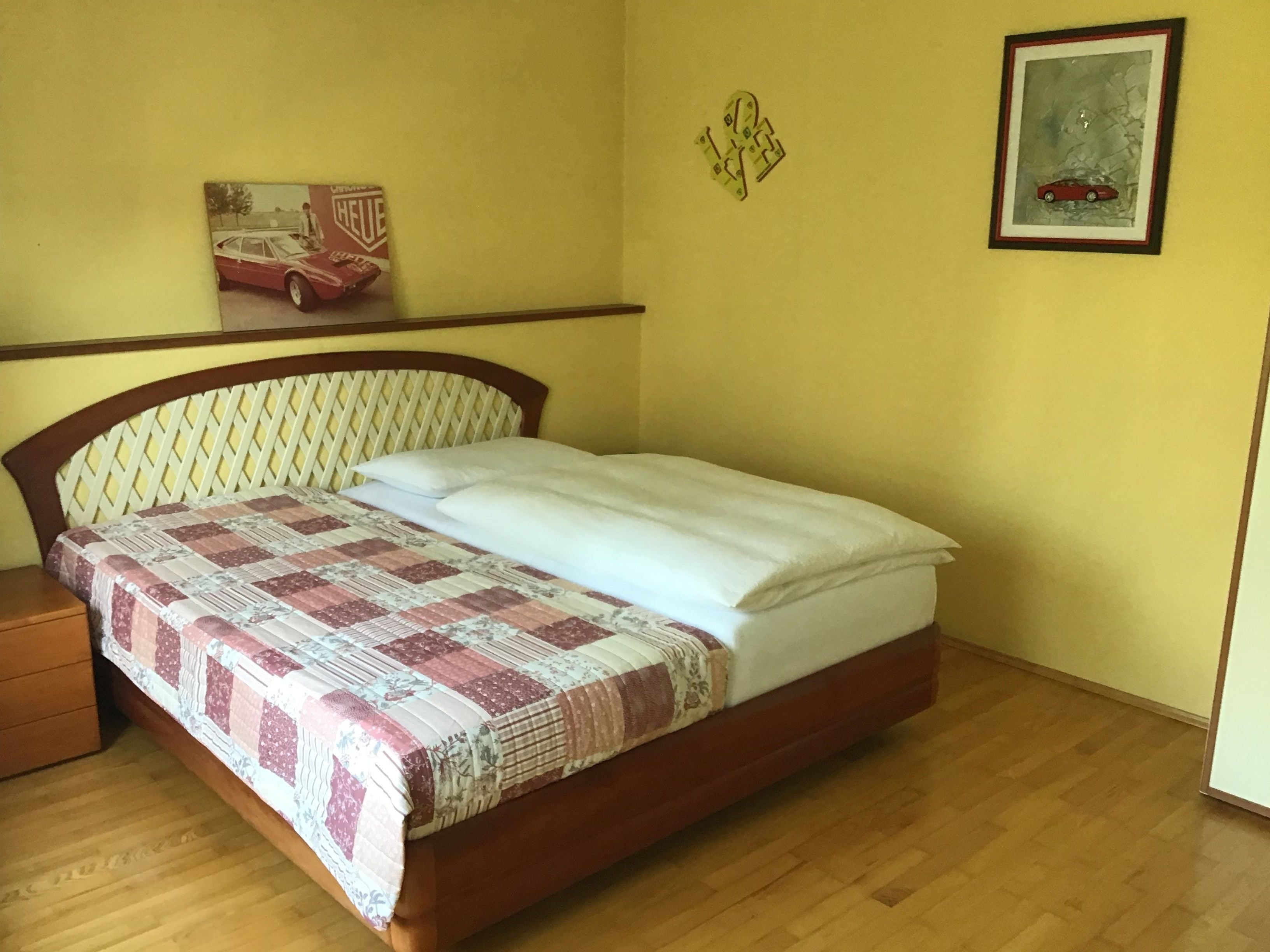 Schlafzimmer mit Bett, kariertem Bettüberwurf, gelben Wänden und Auto-Bildern an der Wand.