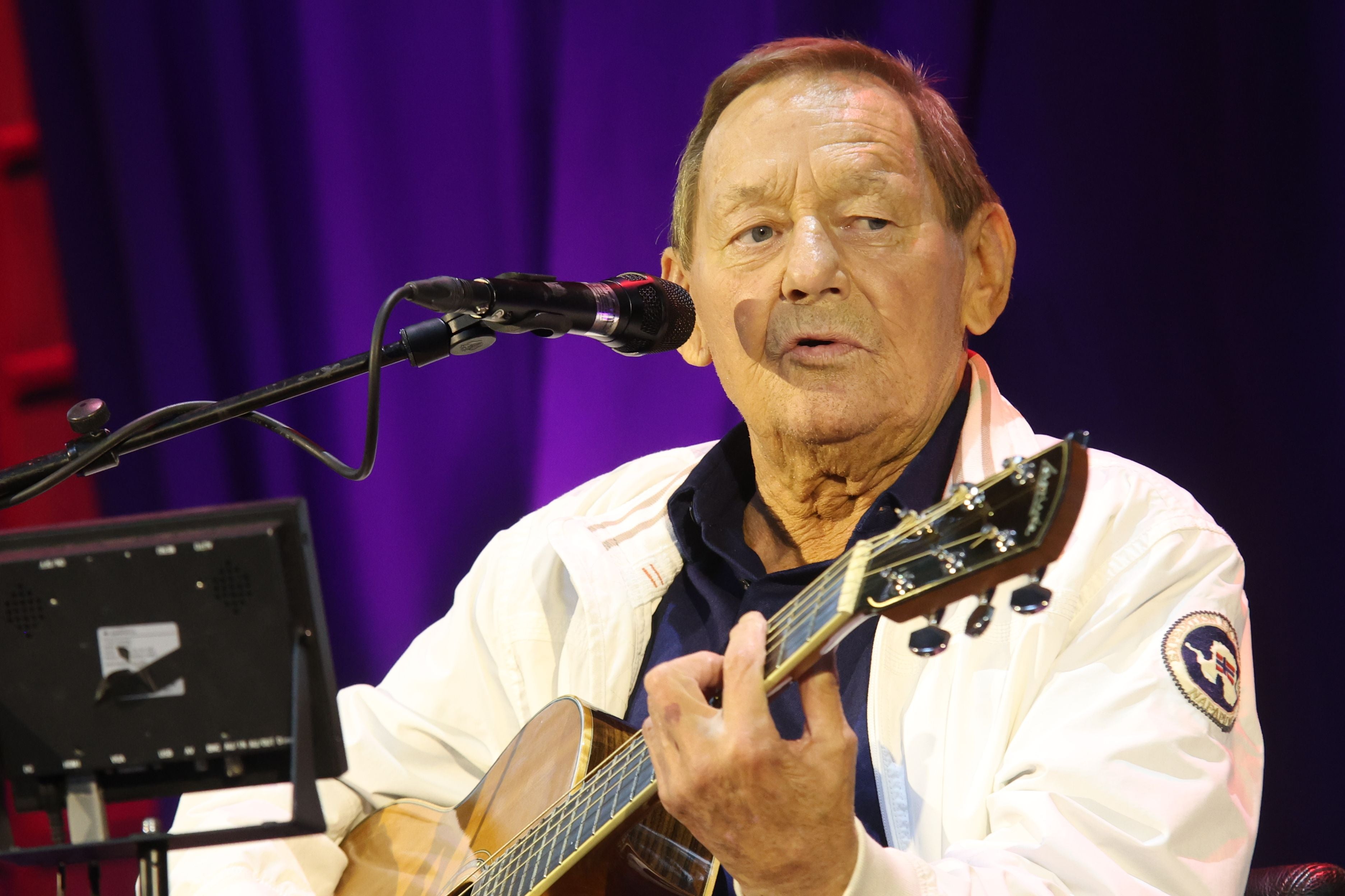Ein älterer Mann spielt Gitarre und singt auf einer Bühne mit violettem Hintergrund.