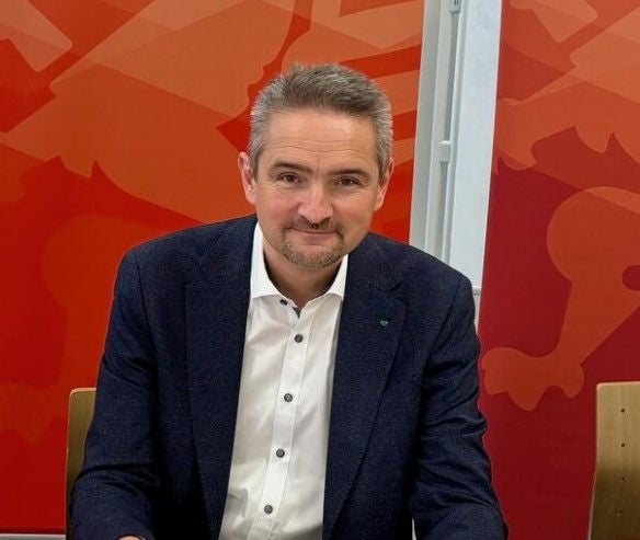 Ein Mann im Anzug sitzt vor einer roten Wand und lächelt in die Kamera.