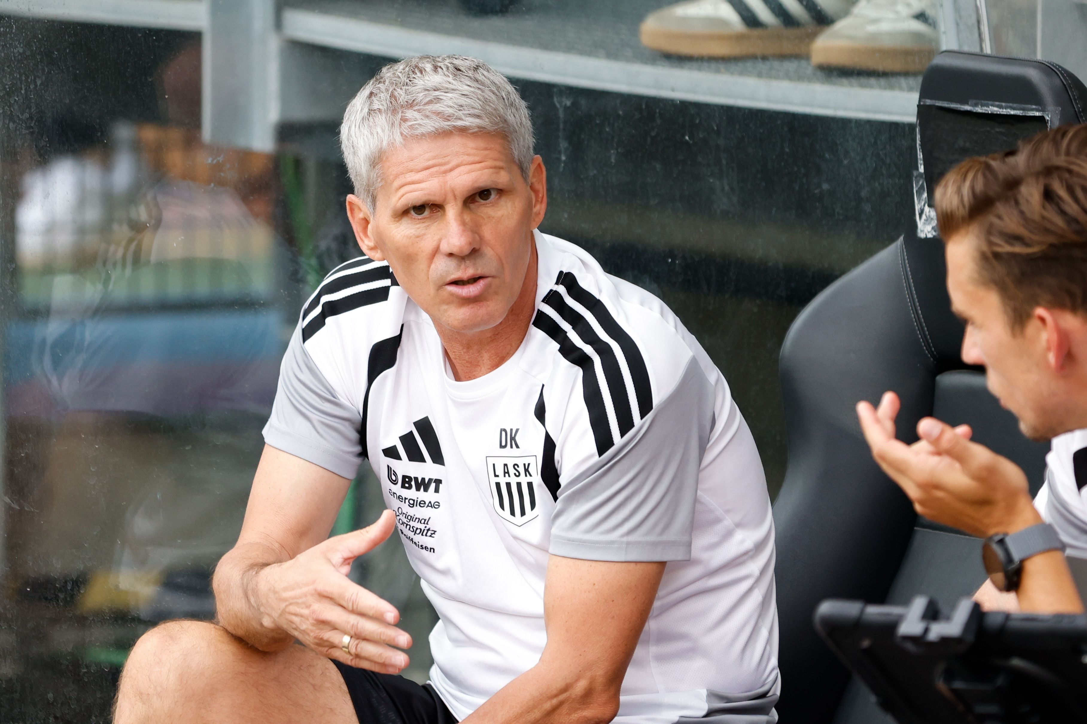 Ein Fußballtrainer im LASK-Trikot spricht auf der Ersatzbank mit einem Kollegen.