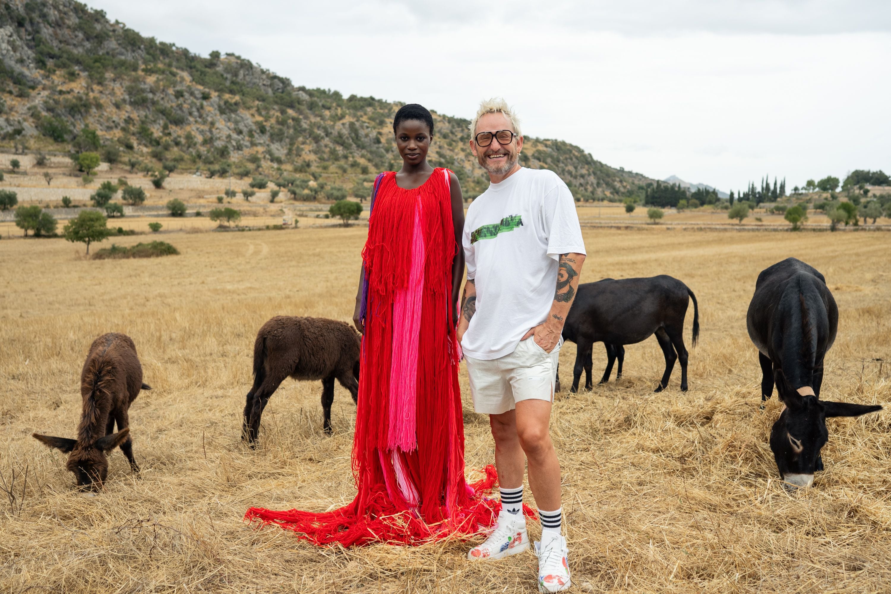 Eine Frau im roten Kleid und ein Mann stehen auf einem Feld mit mehreren grasenden Eseln.