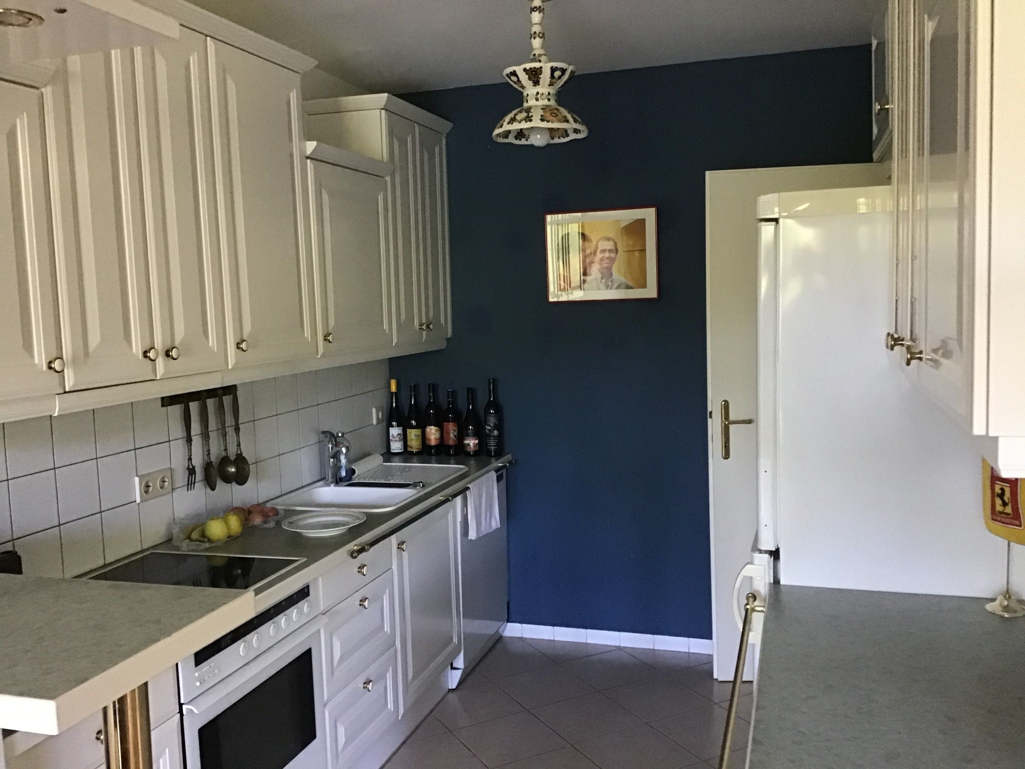 Küche mit weißen Schränken, blauer Wand, Weinflaschen und einem Porträt an der Wand.