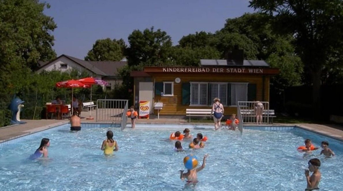 Kinder spielen im Freibad mit Schwimmflügeln, Erwachsene beobachten vom Beckenrand.