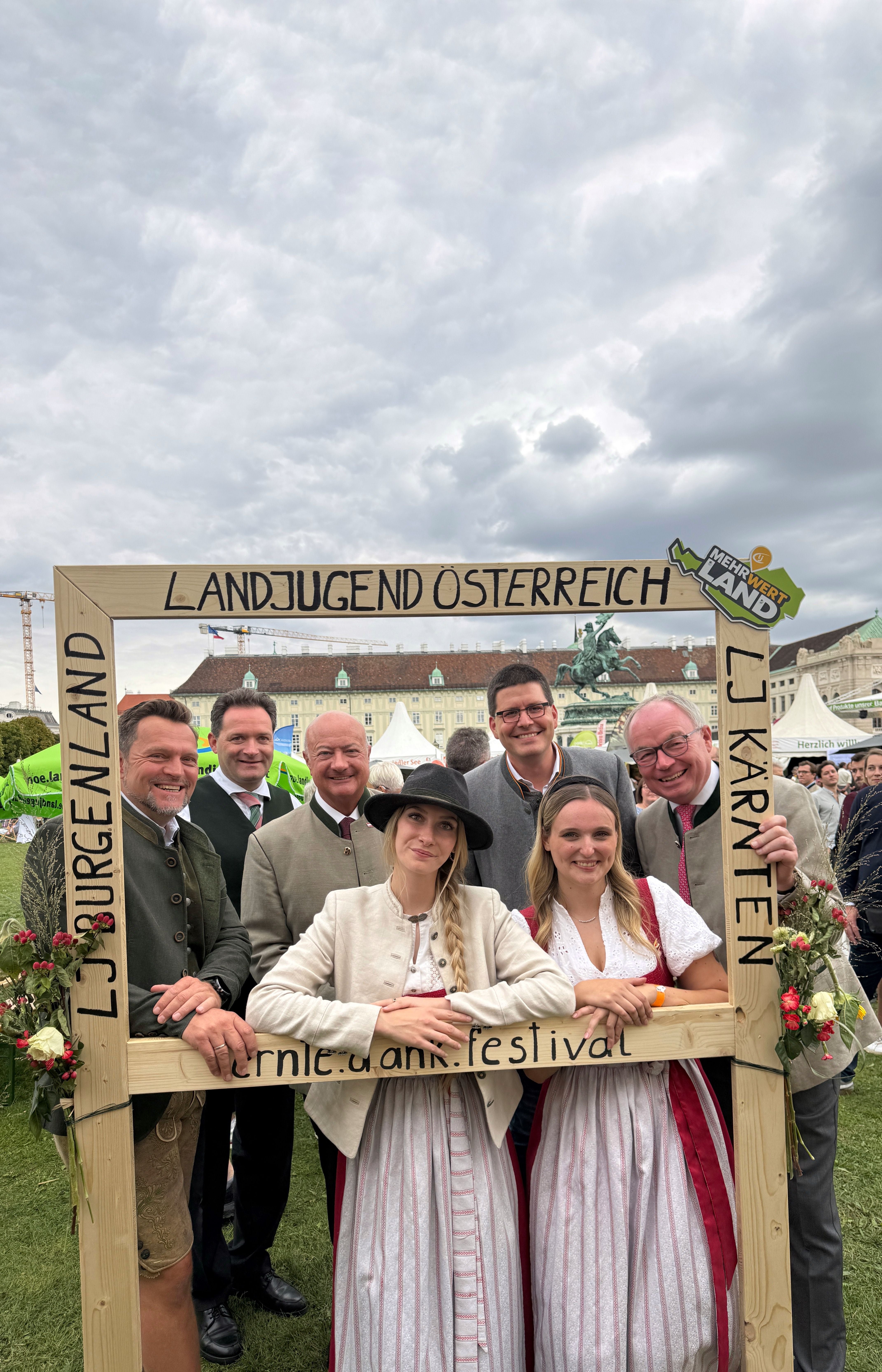 Sieben Personen stehen lächelnd in traditioneller Tracht hinter einem Holzrahmen beim Erntedankfest.
