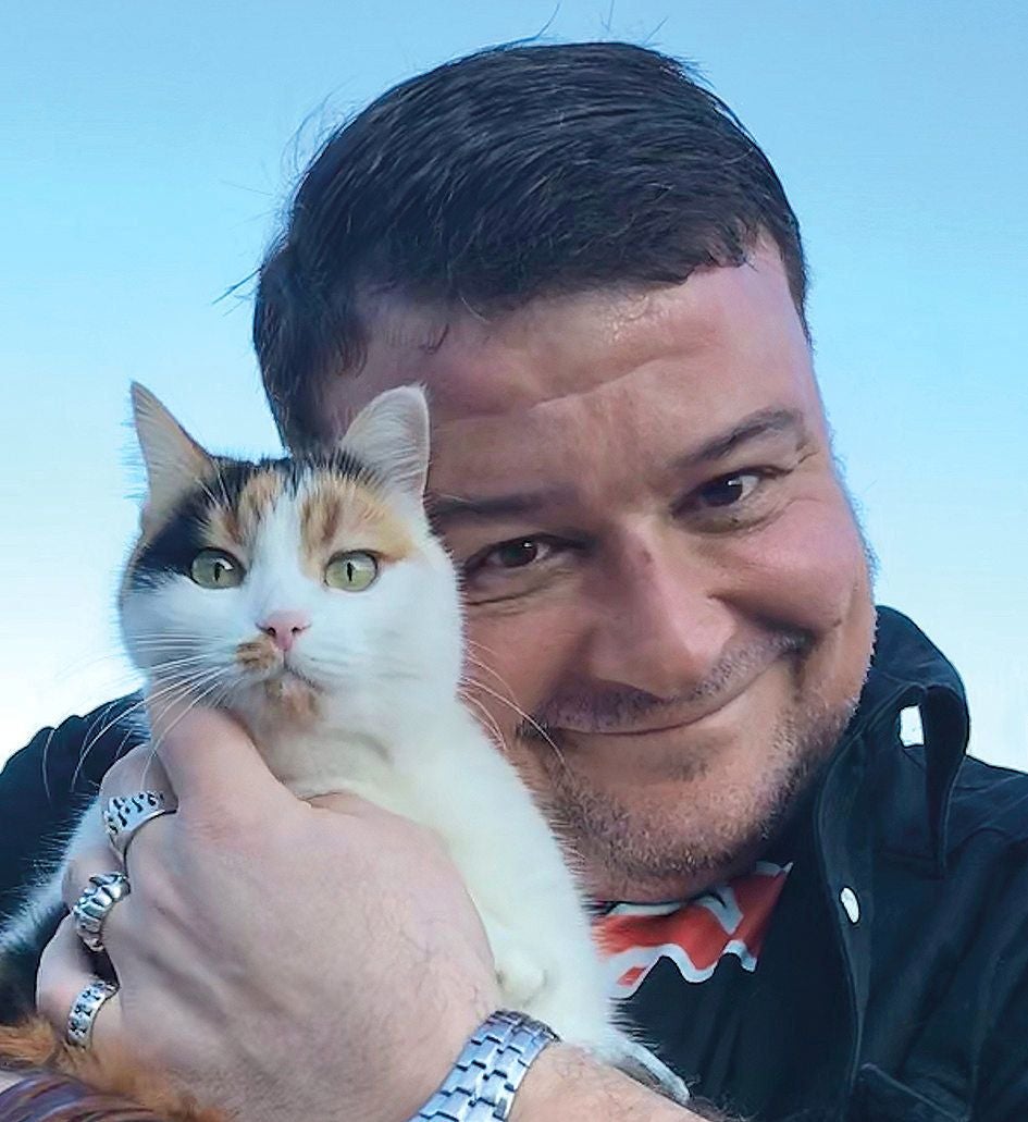 Ein Mann hält eine dreifarbige Katze dicht an sich und lächelt in die Kamera.