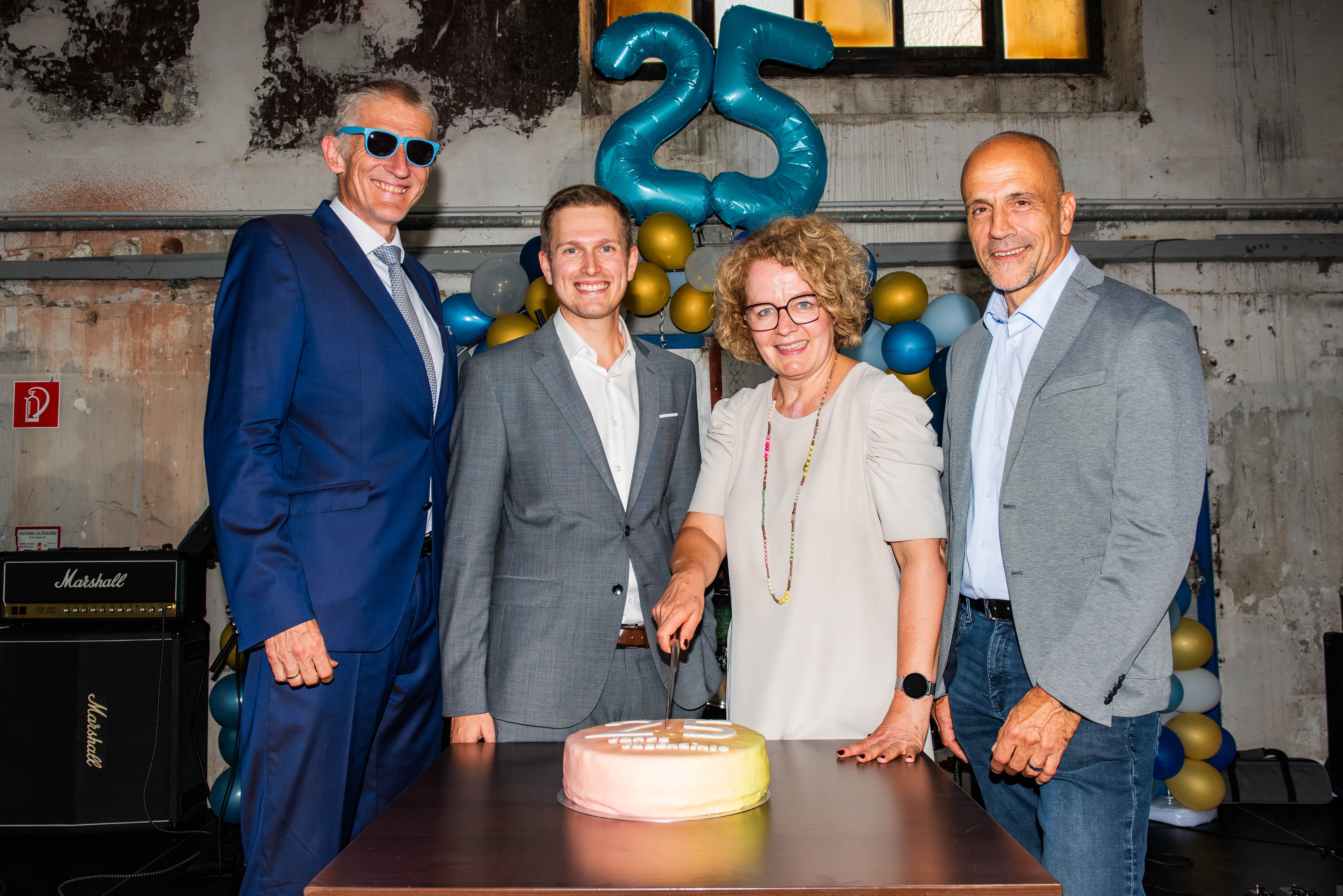 Vier Personen stehen um eine Torte, im Hintergrund sind Ballons mit der Zahl 25 zu sehen.