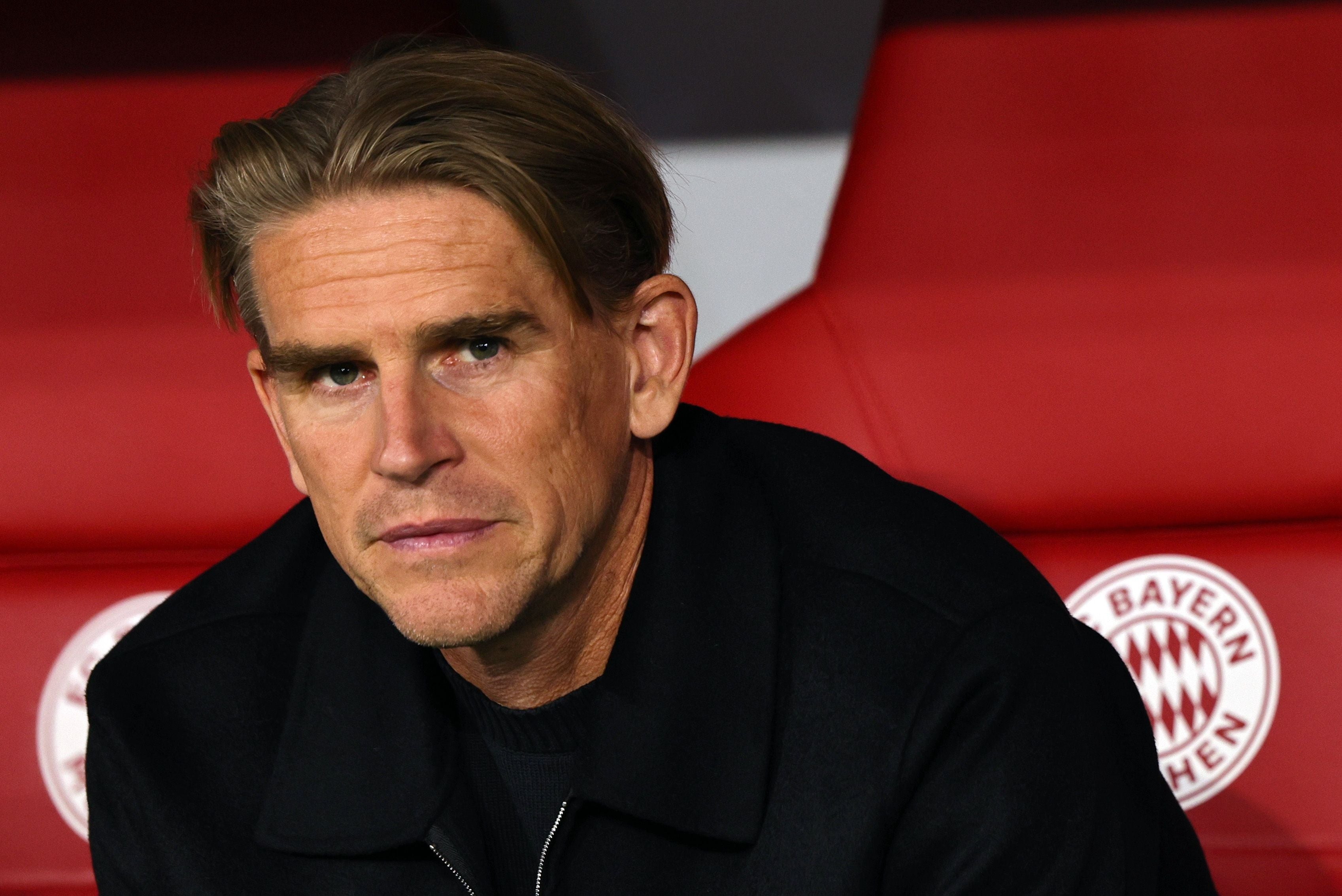 Bayern-Sportdirektor Christoph Freund sitzt im dunklen Oberteil vor der roten Trainerbank mit Bayern-München-Logo und blickt aufmerksam zur Seite.
