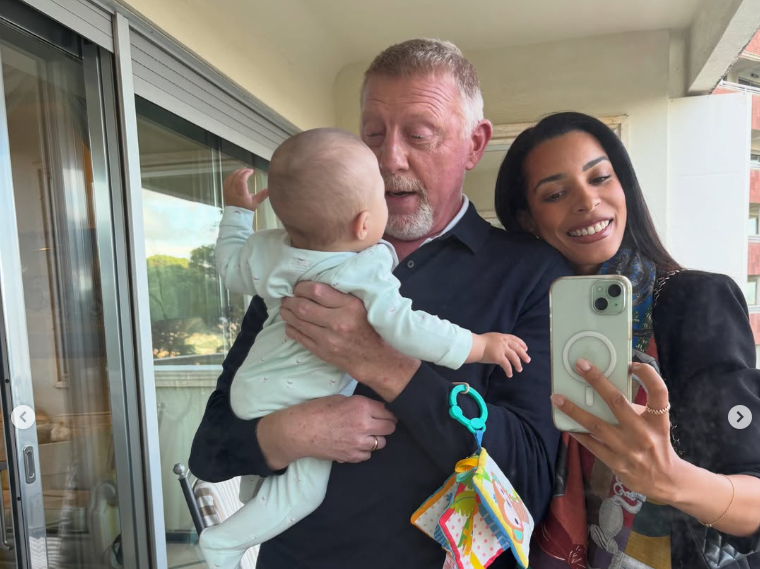 Ein älterer Mann hält ein Baby, eine Frau macht ein Selfie mit ihnen und lächelt.