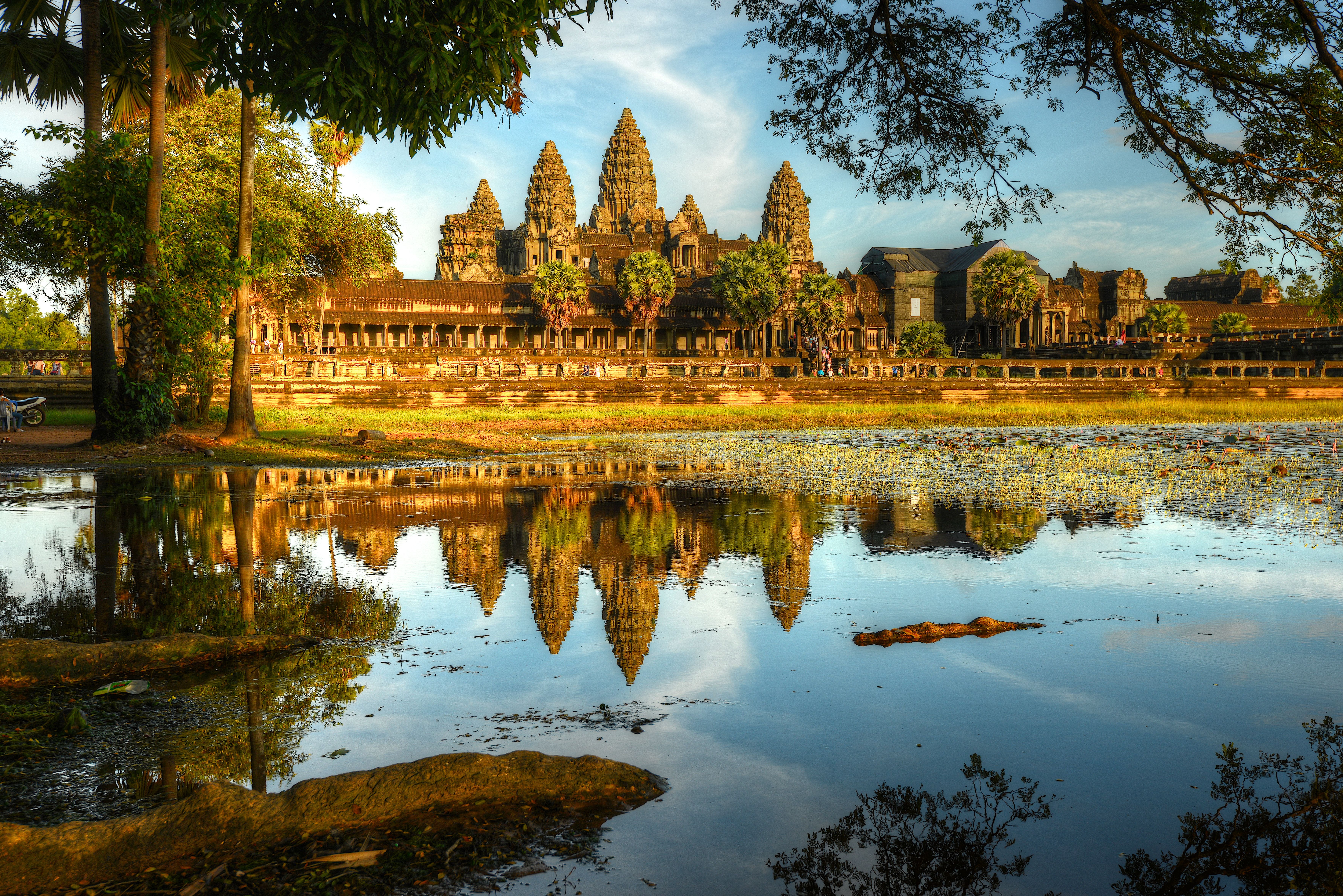 Angkor Wat spiegelt sich im Wasser, umgeben von Bäumen und blauem Himmel.