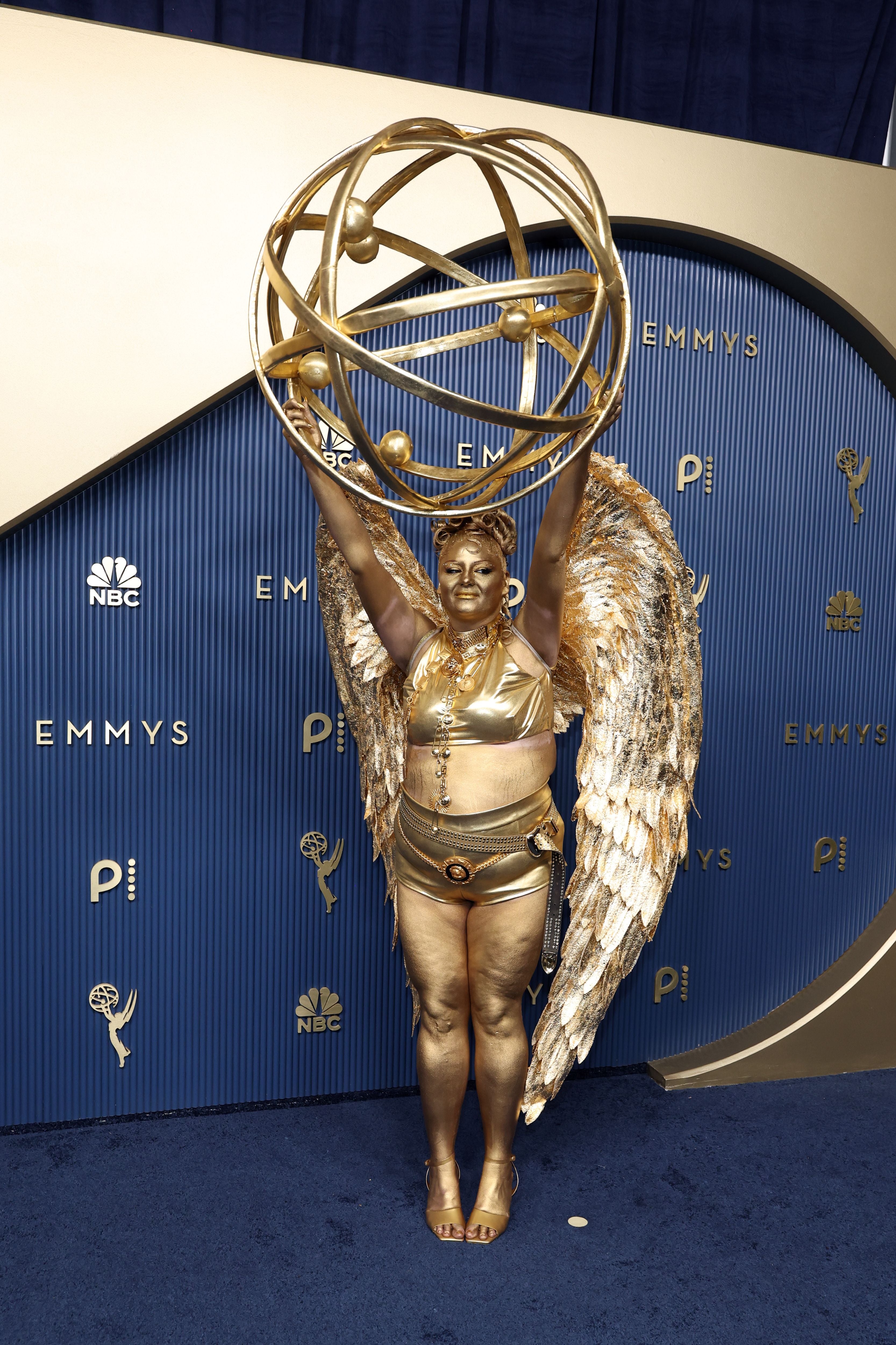Person in goldfarbenem Kostüm mit Engelsflügeln und großer Kugel bei den Emmy Awards.
