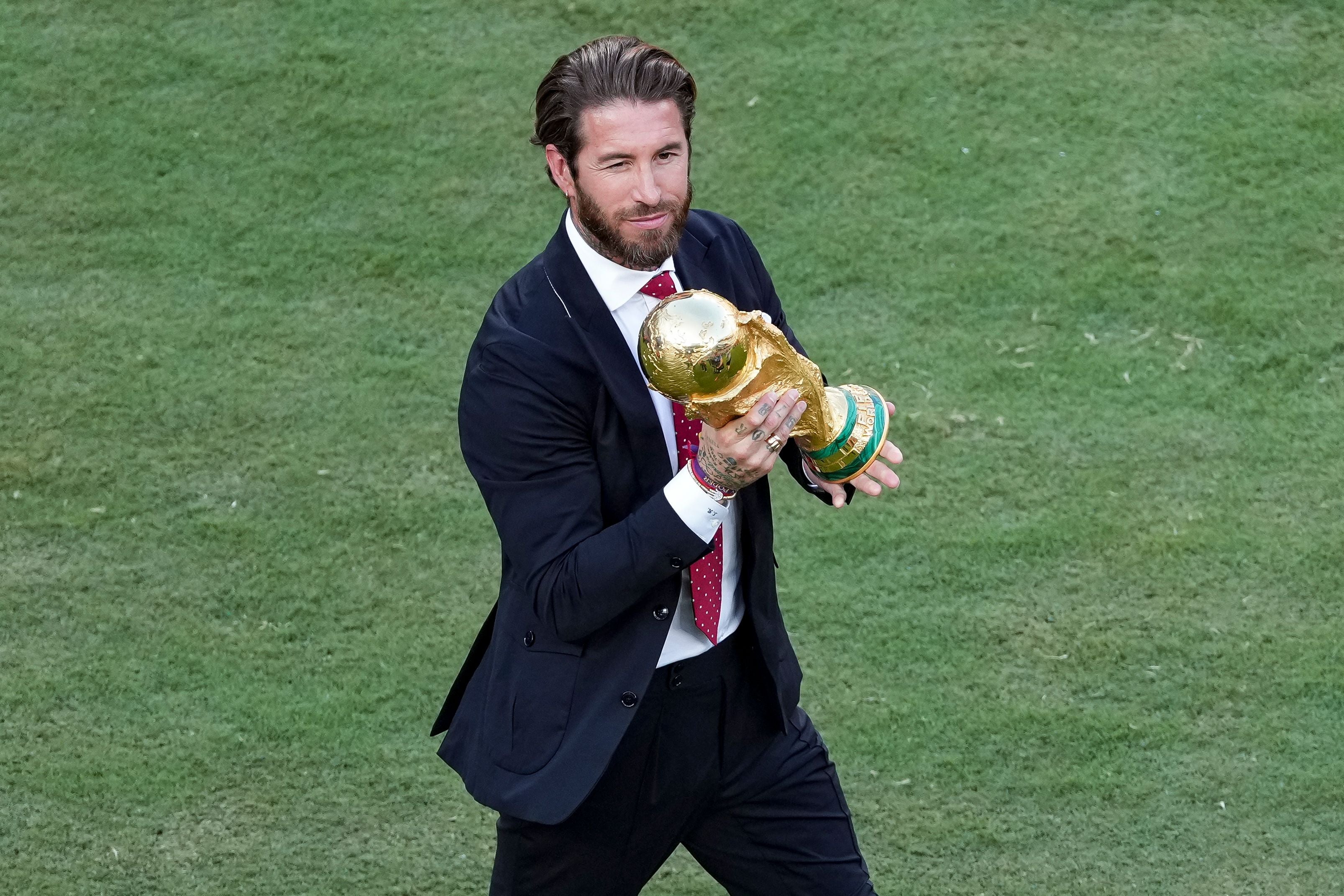 Ein Mann im Anzug hält den WM-Pokal und zwinkert auf einem Fußballfeld.
