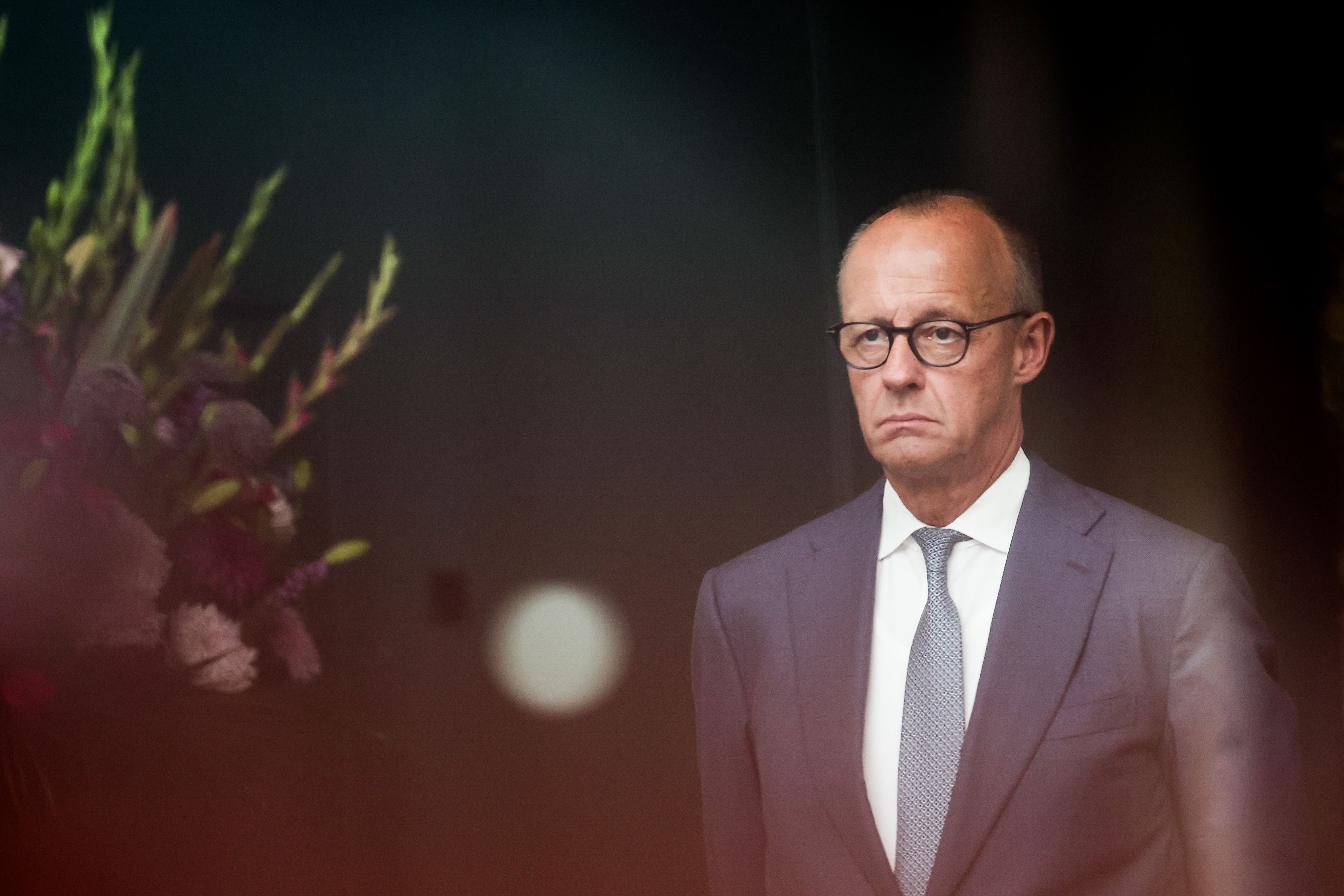 Ein Mann im Anzug mit ernster Miene steht vor dunklem Hintergrund und Blumenarrangement.