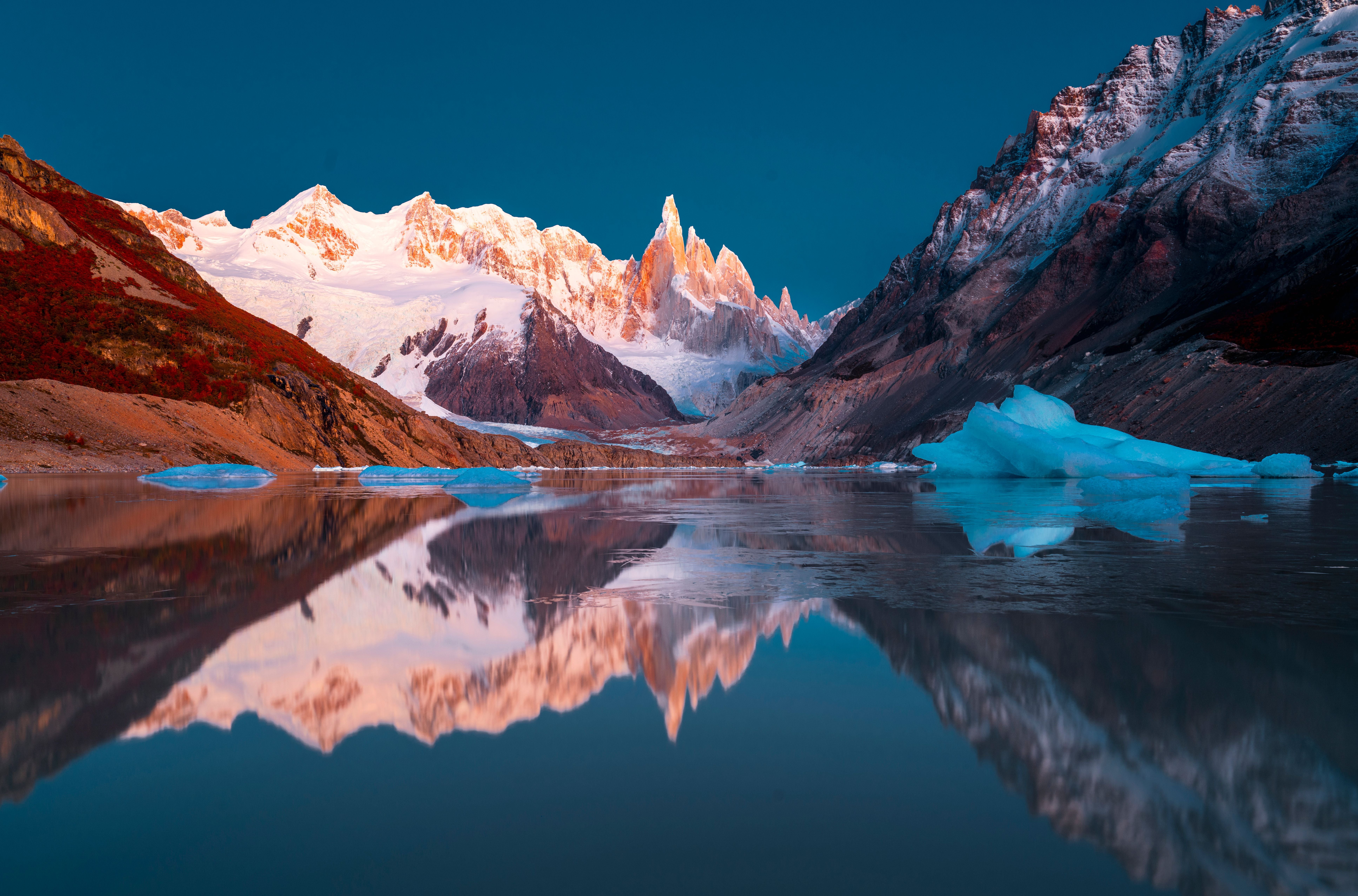 Schneebedeckte Berge spiegeln sich im ruhigen See mit schwimmenden Eisblöcken.