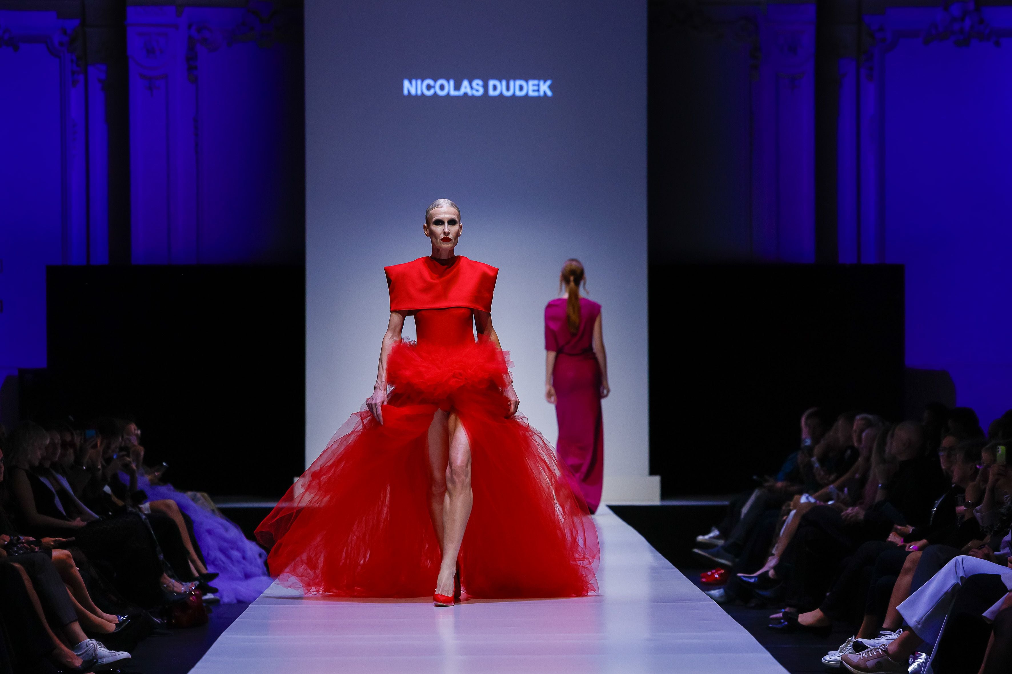 Model in rotem Tüllkleid läuft auf dem Laufsteg, Publikum sitzt an den Seiten.