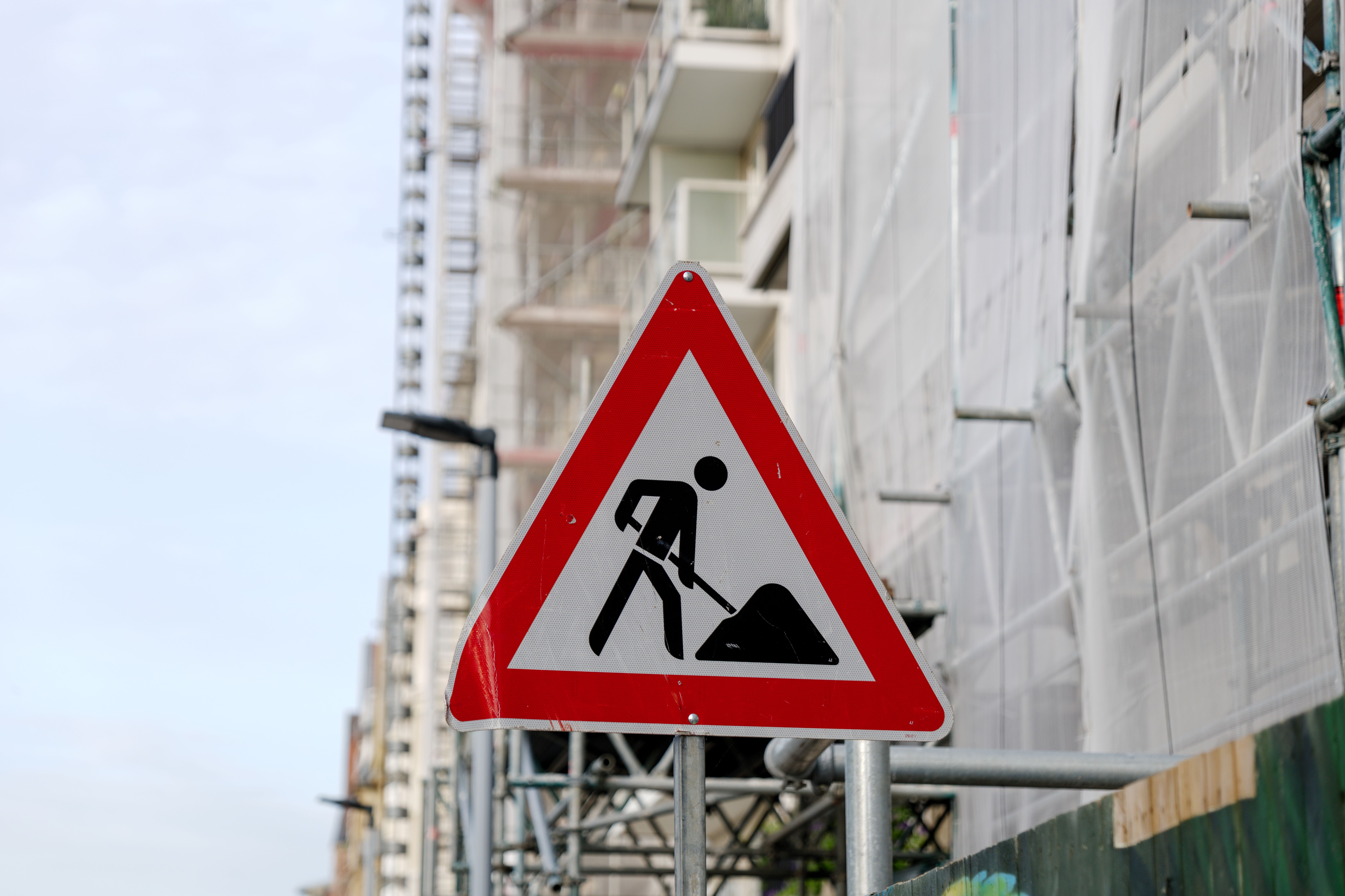 Warnschild für Baustelle auf einer Straße vor einem Gebäude mit Baugerüst im Hintergrund.