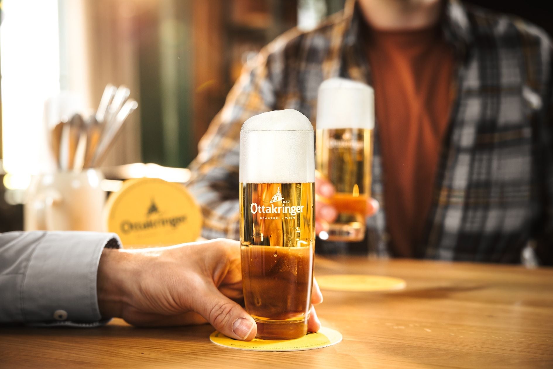 Zwei Personen stoßen mit Ottakringer Biergläsern an einem Holztisch an.