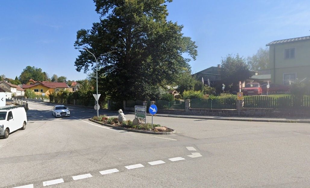 Straßenecke mit Verkehrsinsel, Bäumen, Autos und Wohnhäusern bei sonnigem Wetter.