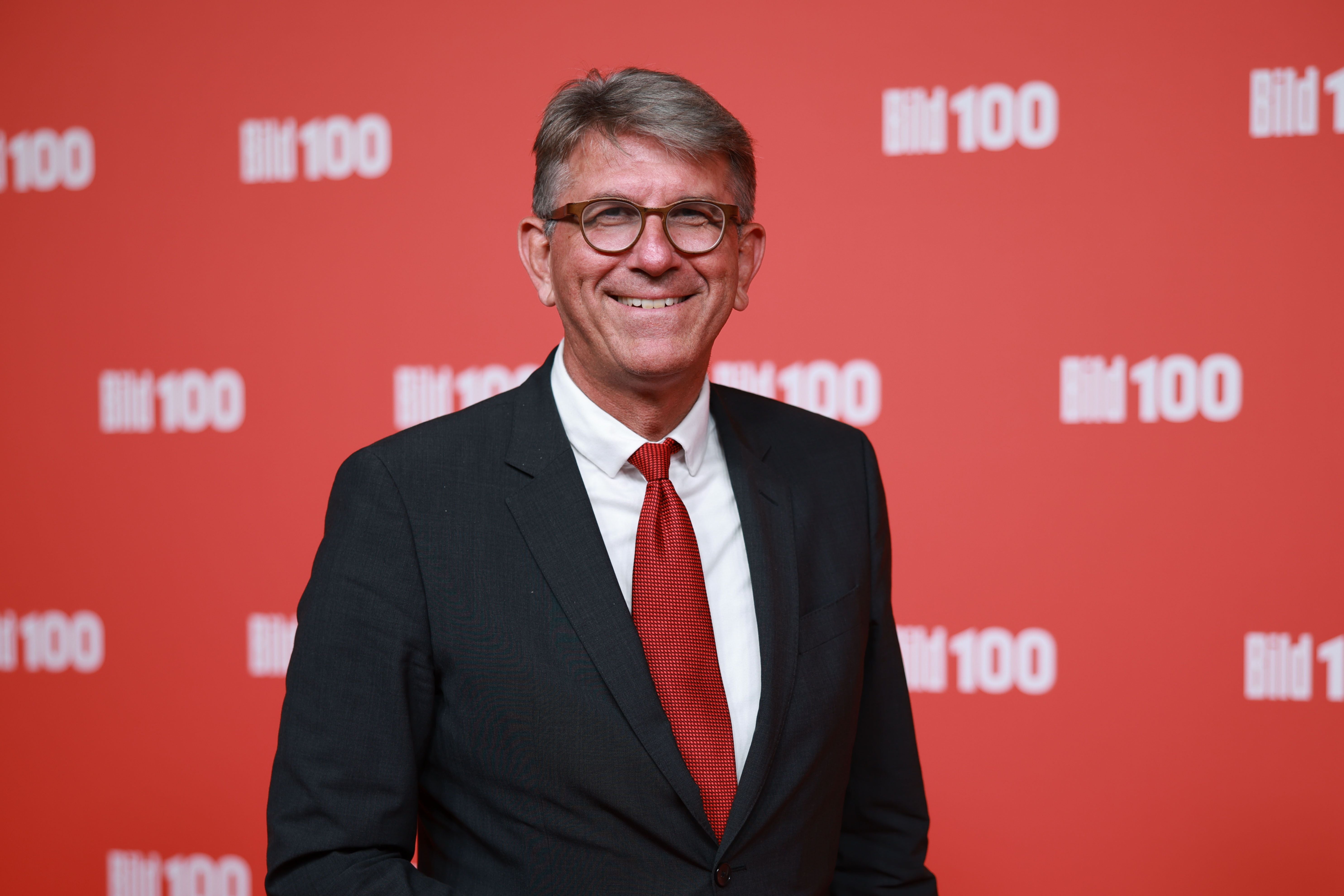 Ein Mann im Anzug mit roter Krawatte steht vor rotem "Bild 100"-Hintergrund und lächelt.