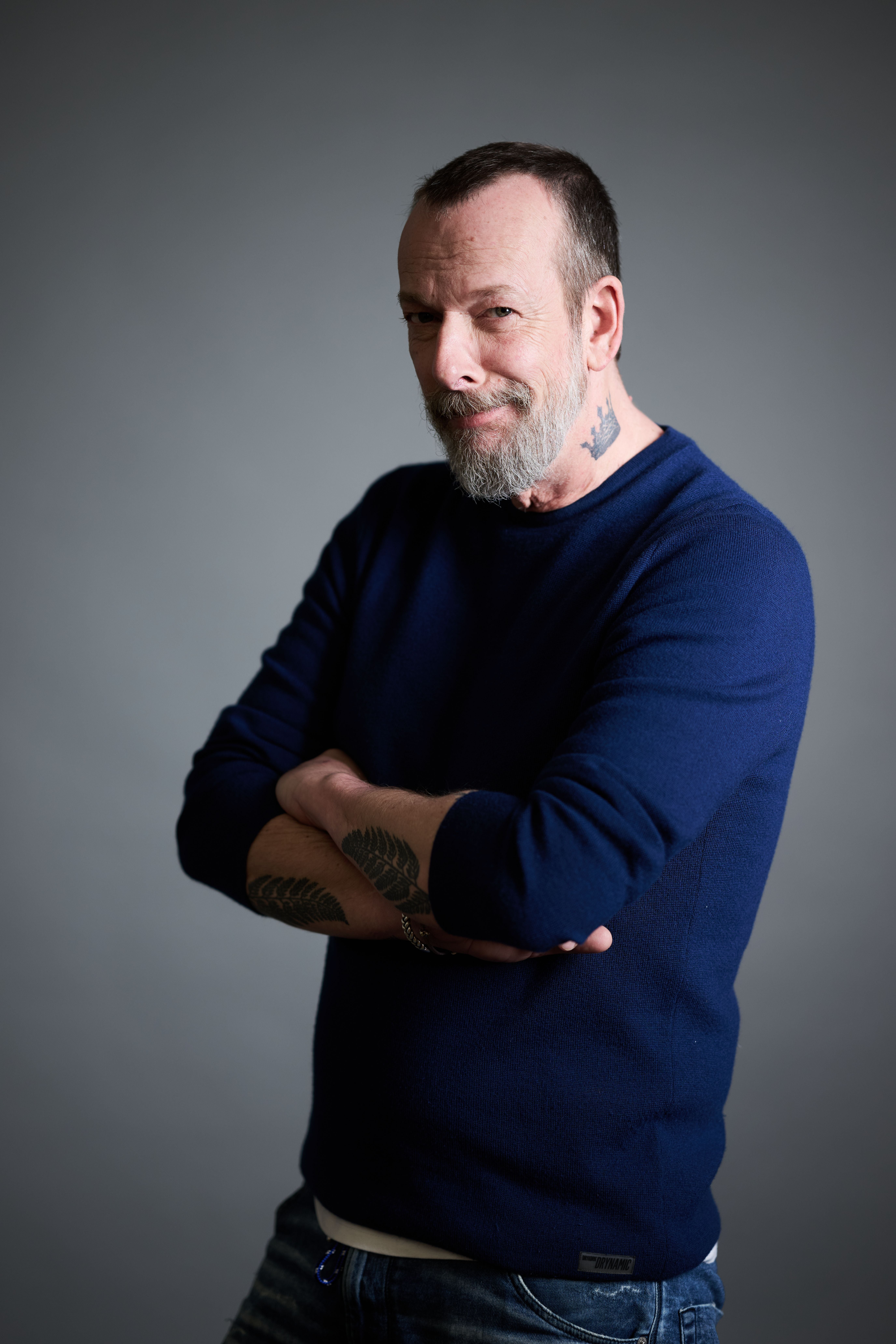 Mann mit Bart und Tattoos, trägt blauen Pullover und verschränkt die Arme, vor grauem Hintergrund.