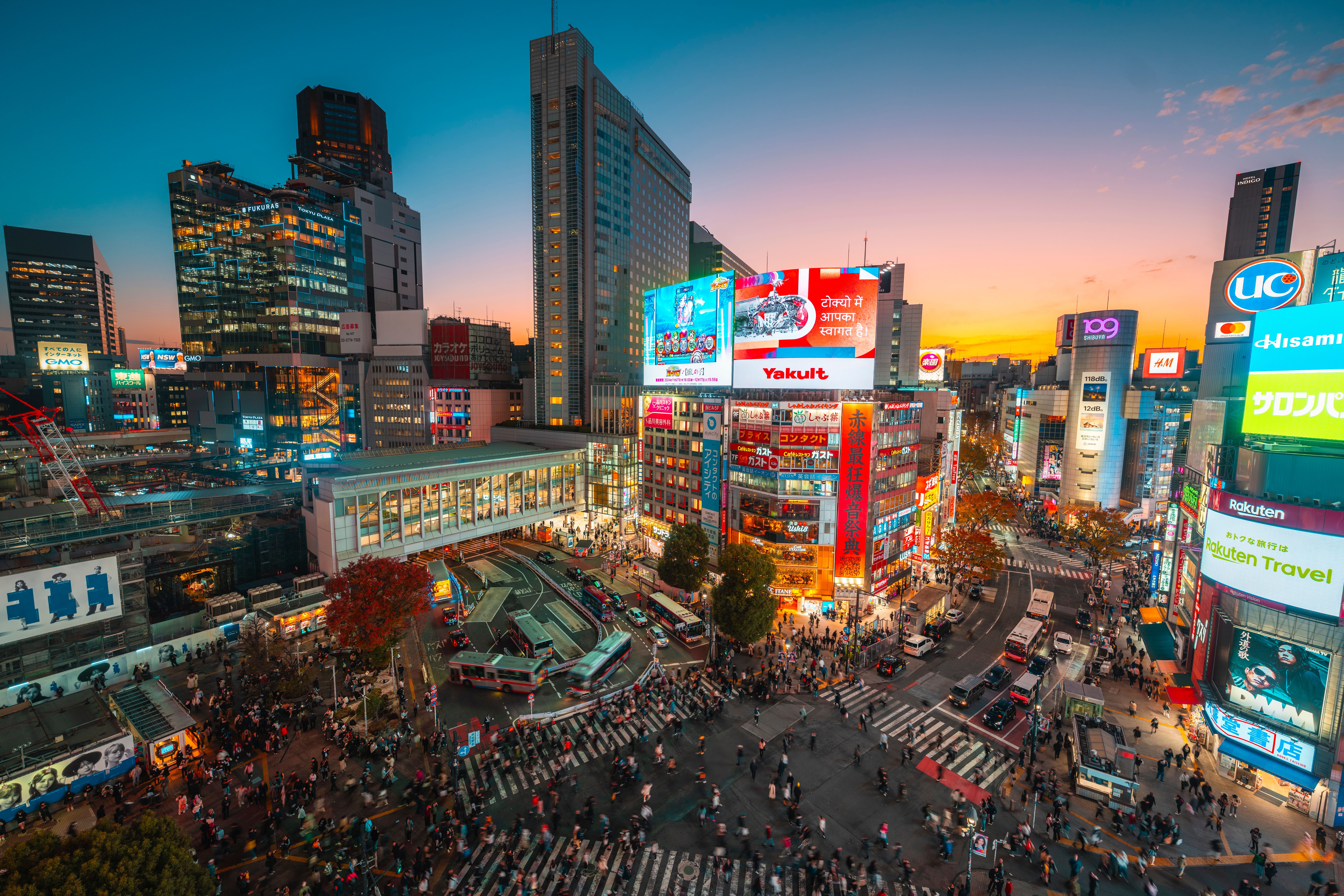 Menschenmengen an einer großen Kreuzung in Tokio bei Sonnenuntergang, umgeben von hohen Gebäuden und Leuchtreklamen.