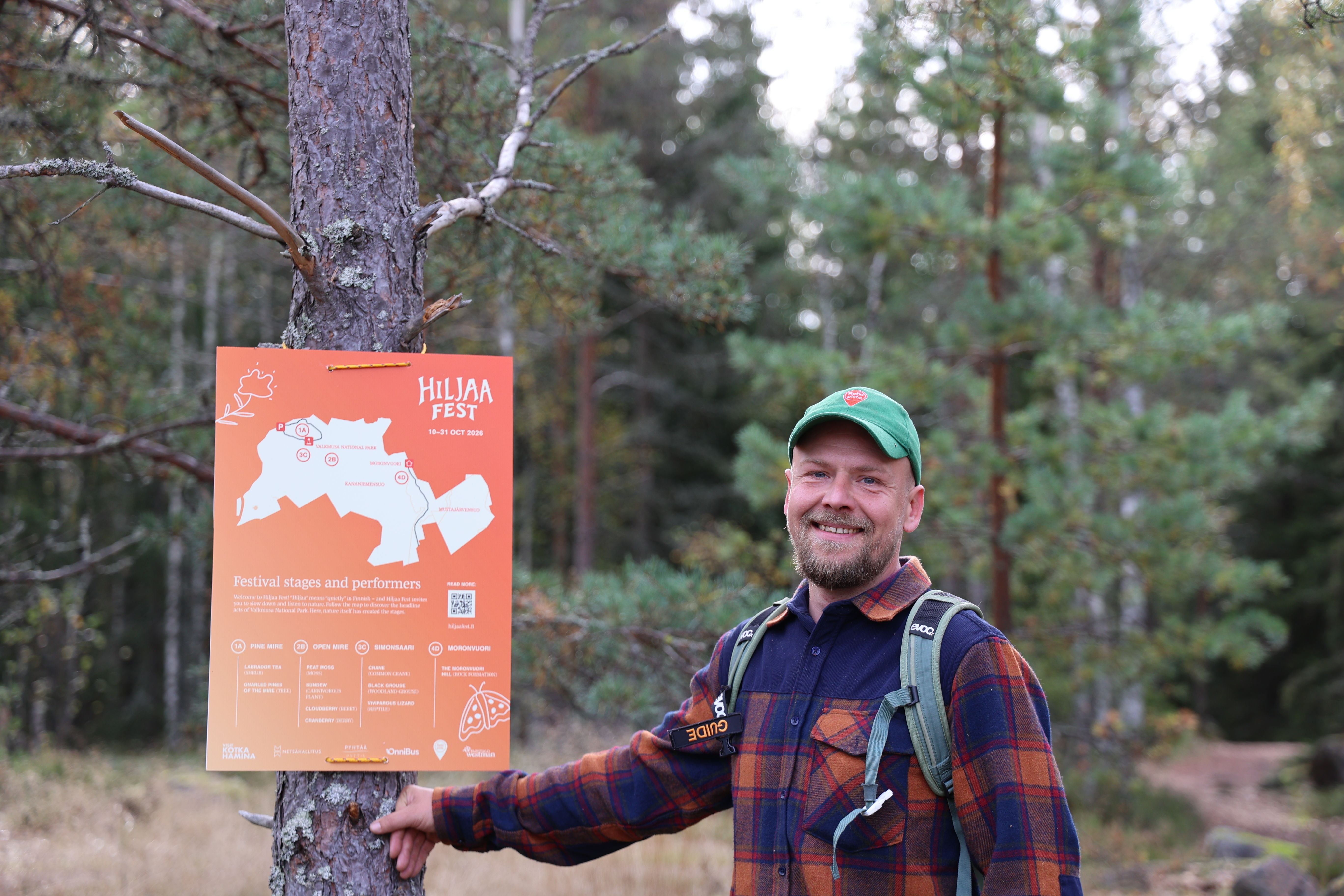 Mann mit grünem Cap steht lächelnd im Wald neben einem orangefarbenen Hiljaa-Fest-Poster.