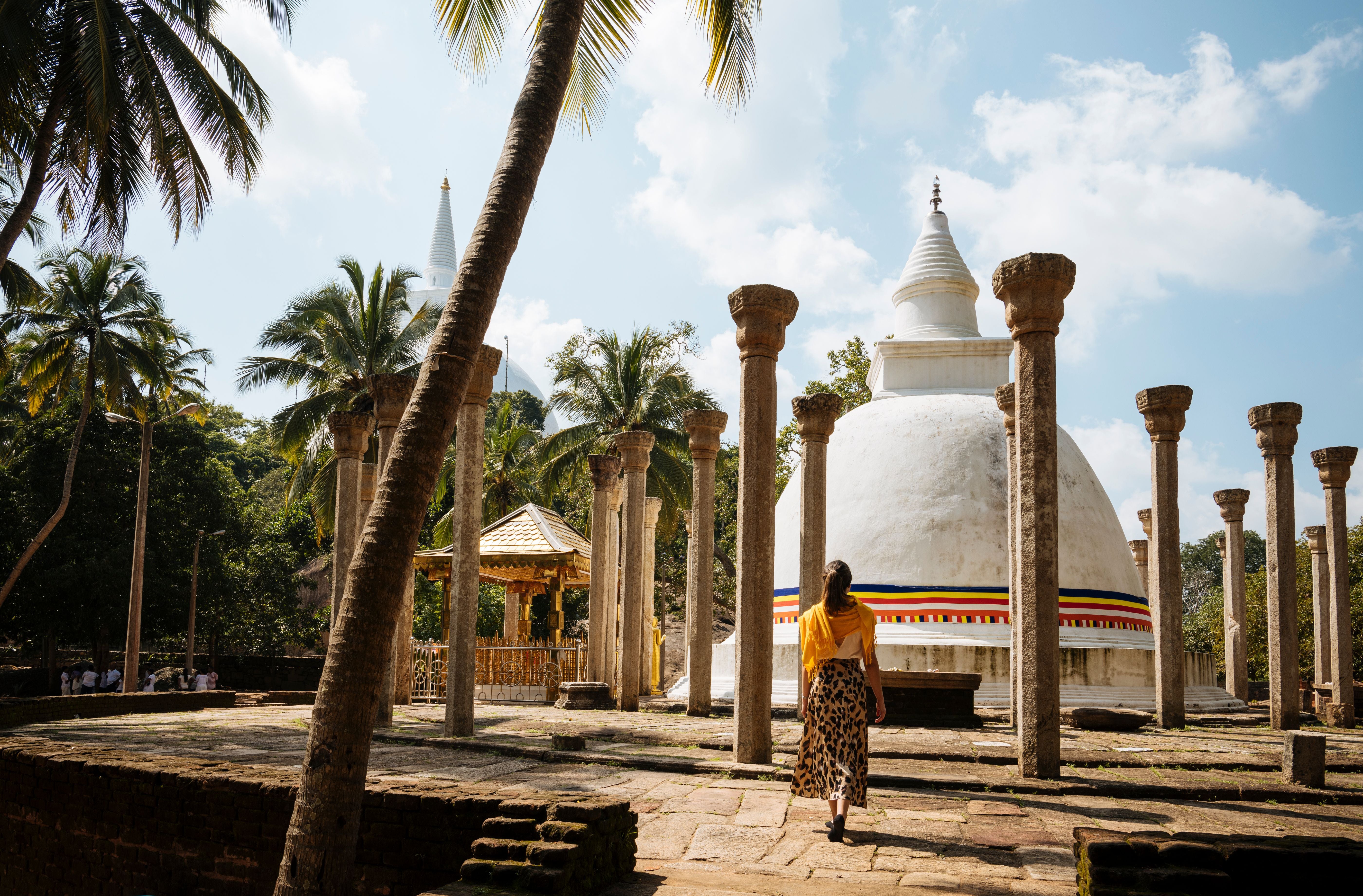 Frau geht zu einem weißen Stupa mit buntem Band, umgeben von Palmen und Säulen in Sri Lanka.