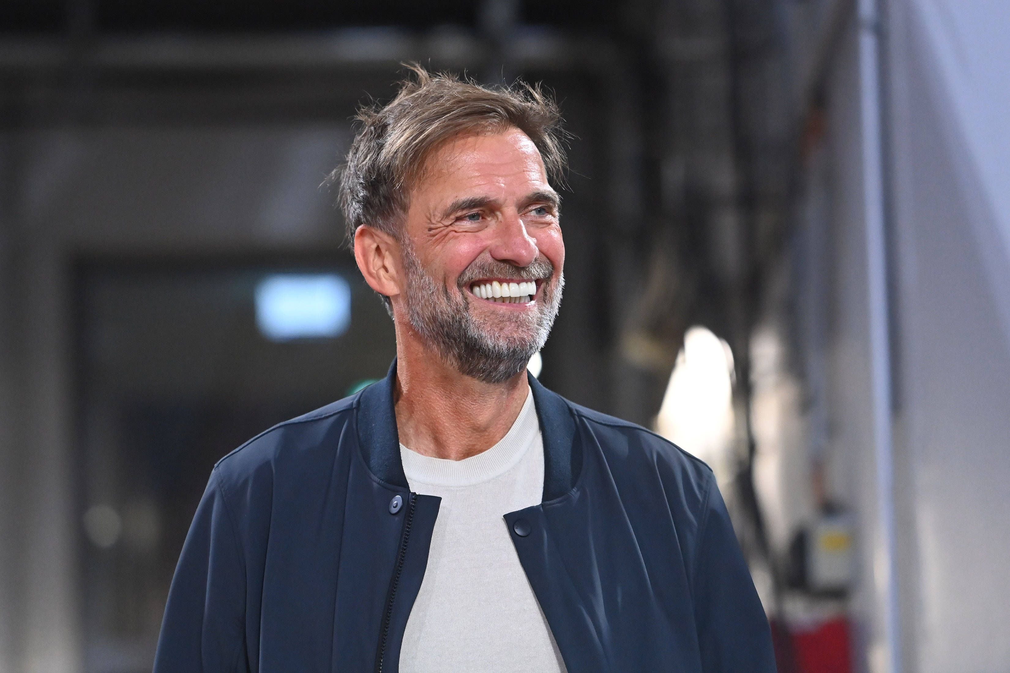 Ein lächelnder Mann mit Bart in heller Kleidung und dunkler Jacke vor unscharfem Hintergrund.