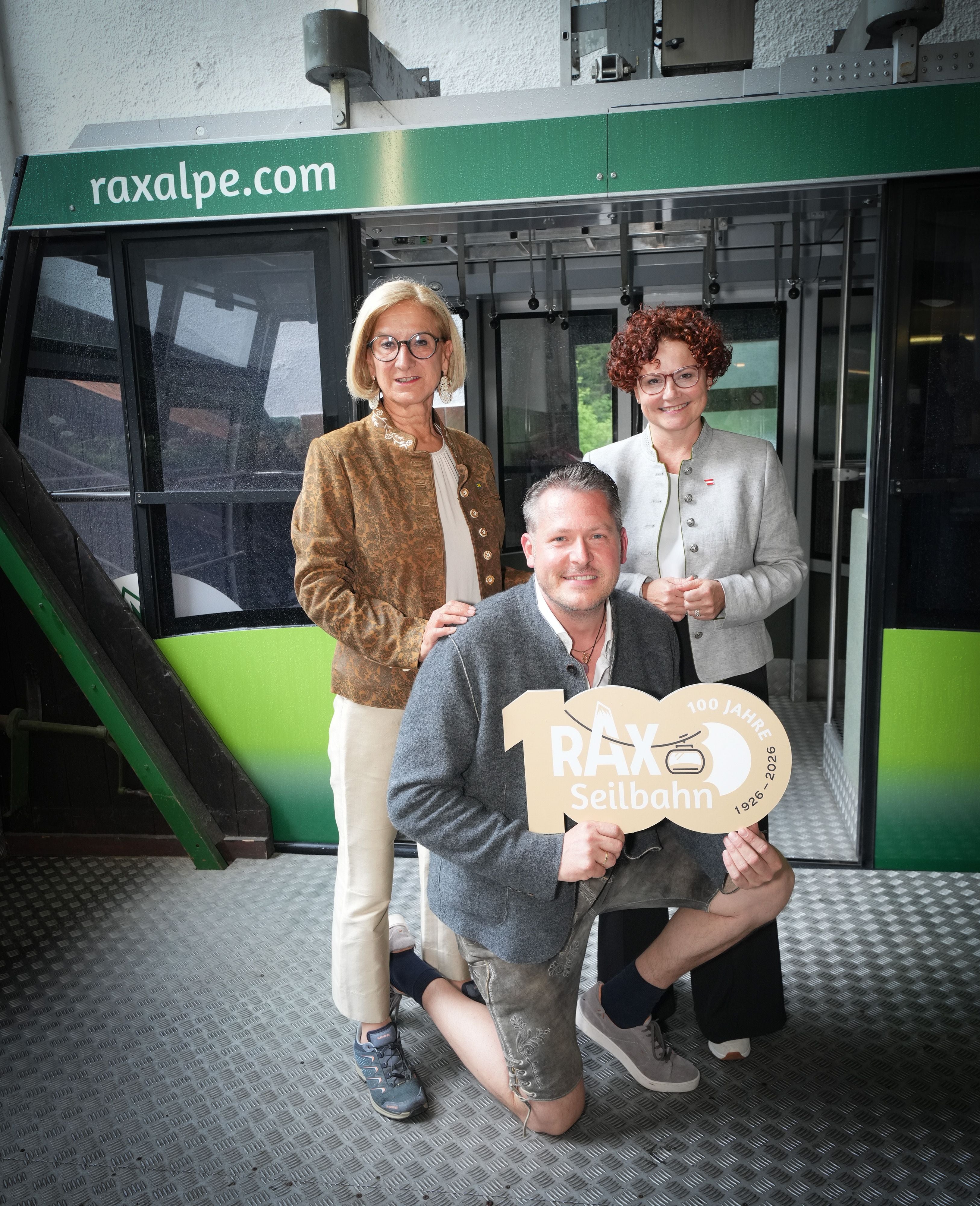 Drei Personen posieren vor einer grünen Rax Seilbahn und halten ein 100-Jahre-Jubiläumsschild.