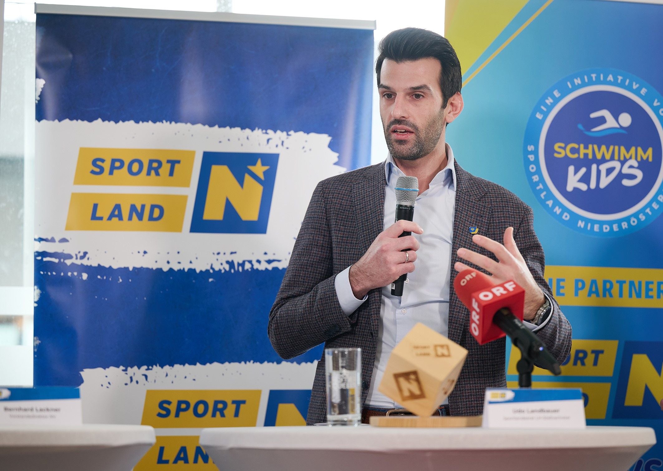Ein Mann spricht bei einer Pressekonferenz mit Mikrophon vor Sportland-Logos.