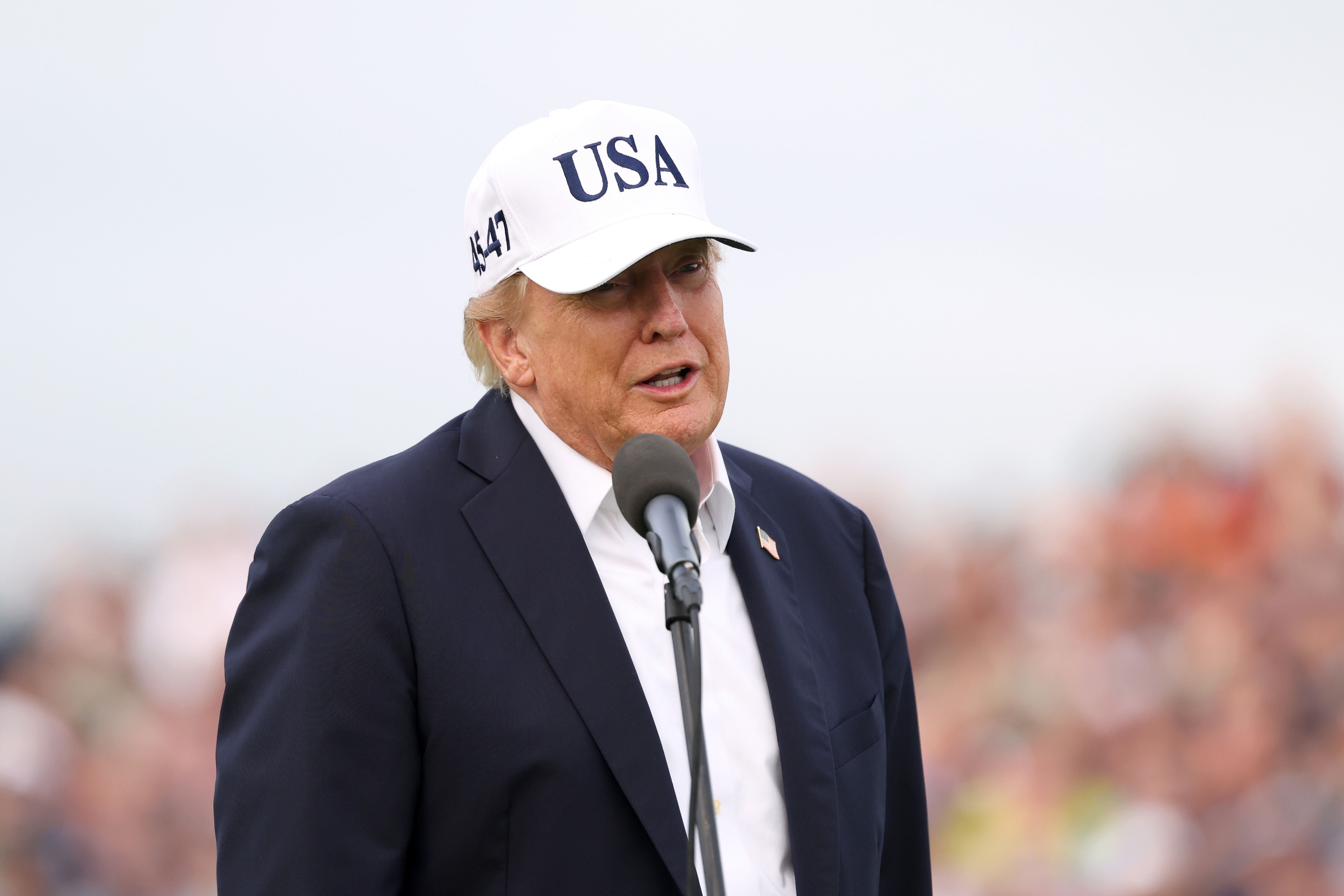 Ein Mann mit weißer "USA"-Kappe spricht draußen in ein Mikrofon vor verschwommenem Hintergrund.