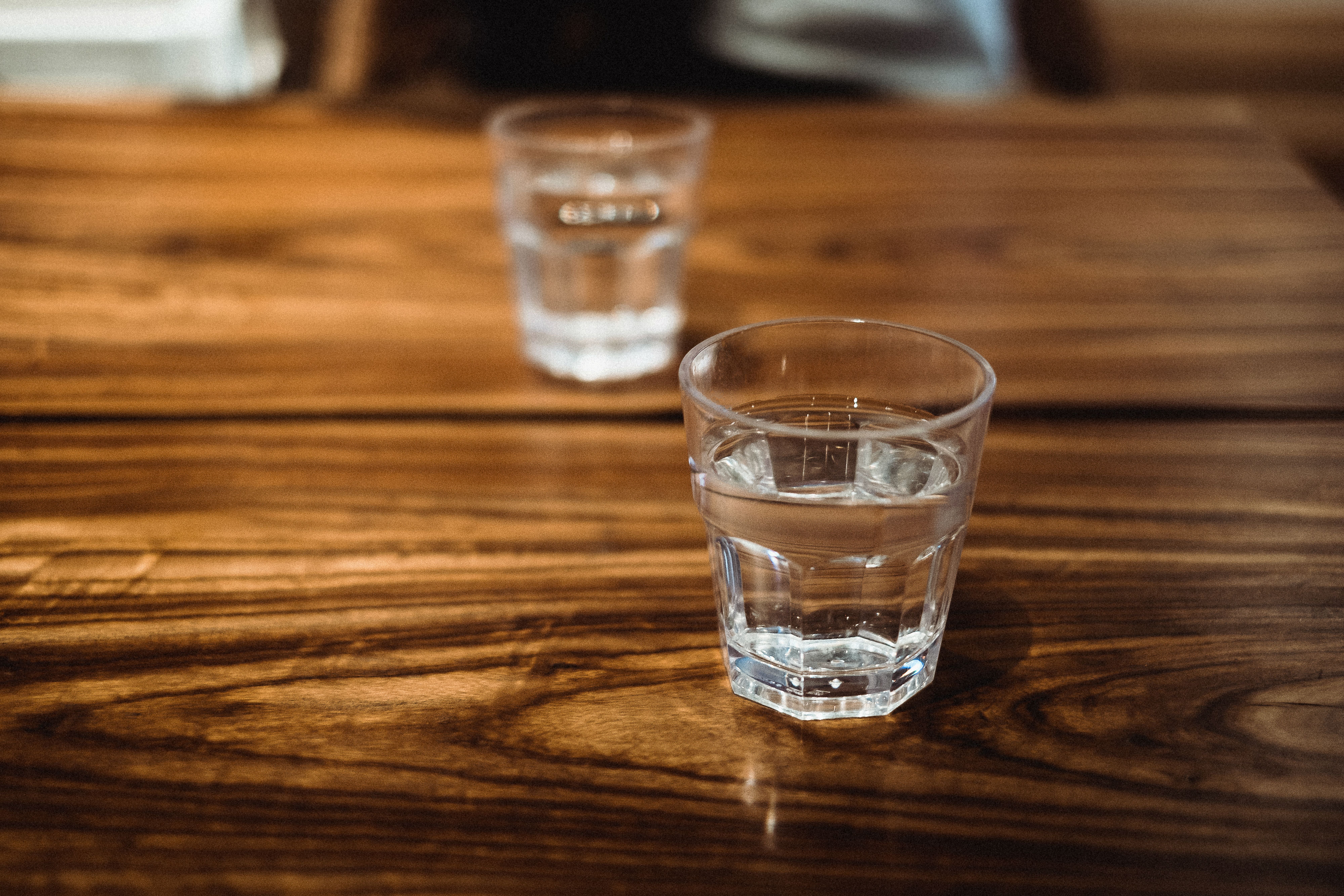 Zwei Wassergläser stehen auf einem Holztisch, eines im Vordergrund, eines im Hintergrund.