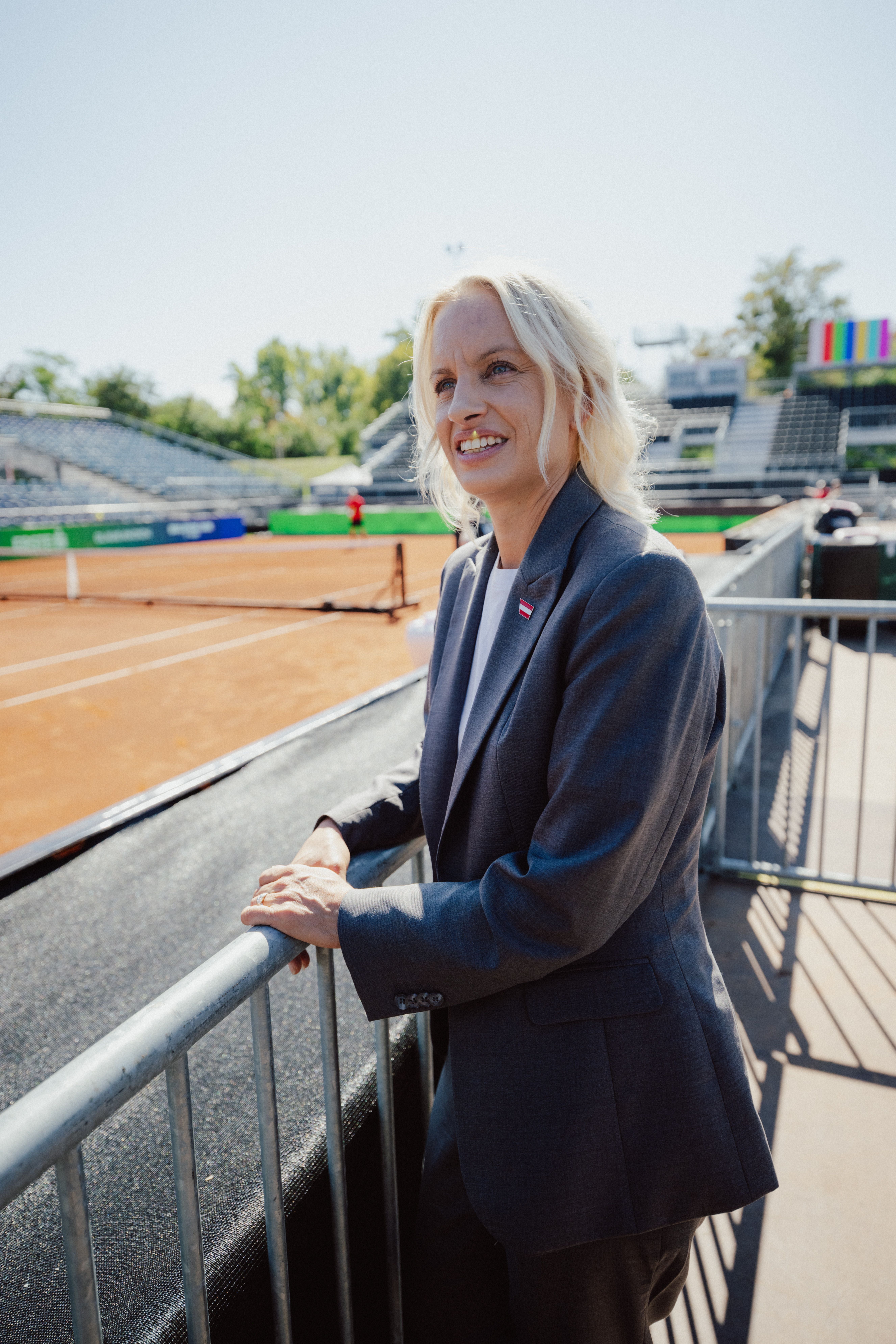 Eine Frau im Anzug steht lächelnd am Rand eines Tennisplatzes im Freien.
