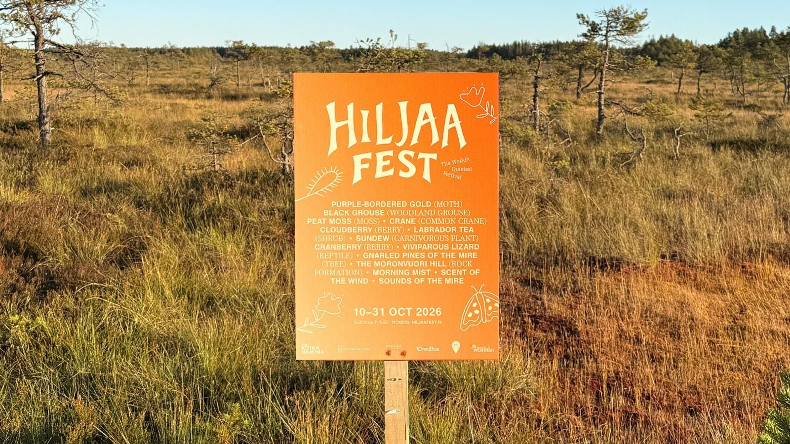 Orange Schild mit "HILJAA FEST" in Moorlandschaft, Bäume und Gras im Hintergrund.