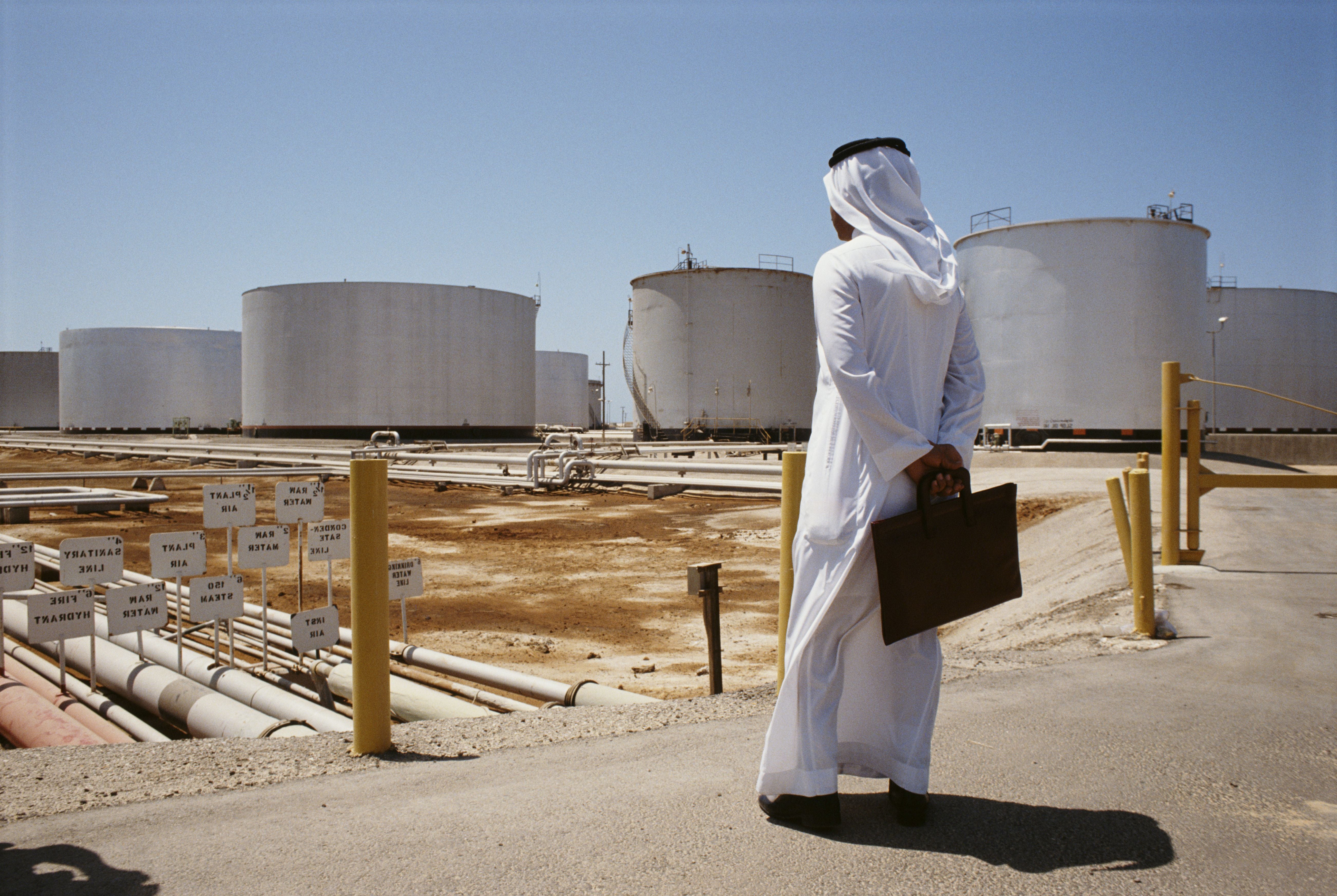 Ein Mann in traditioneller Kleidung mit Aktentasche steht vor großen Öltanks bei Saudi Aramco.