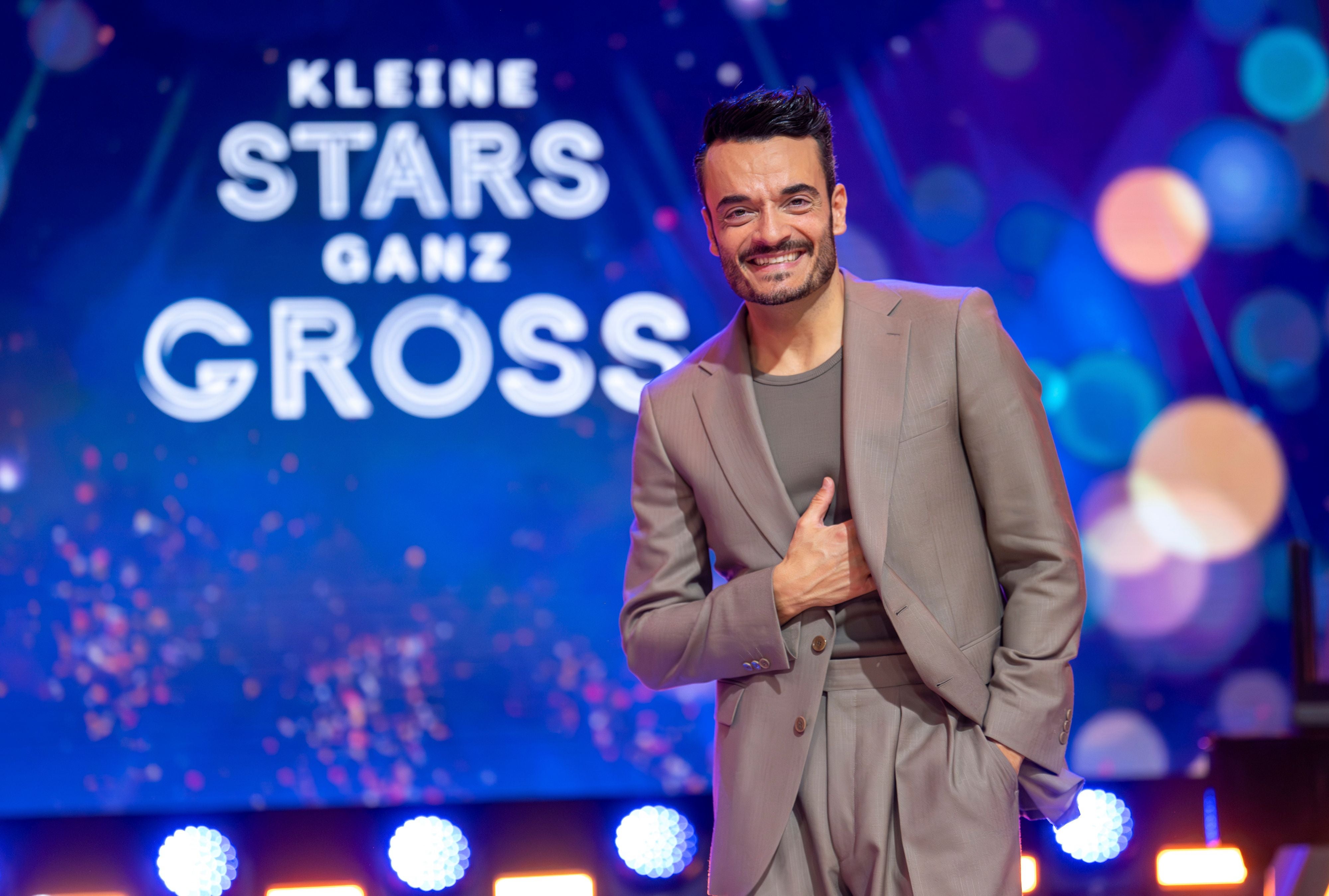 Giovanni Zarrella lächelt im grauen Anzug vor einem „Kleine Stars ganz groß“-Bühnenhintergrund.